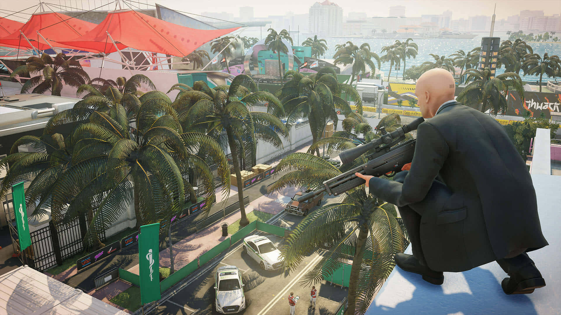 Best Hitman 2 桌布 壁紙 — 一個光頭穿西裝的男人蹲在屋頂上，用狙擊步槍瞄準棕櫚樹對面繁忙的熱帶城市景觀。