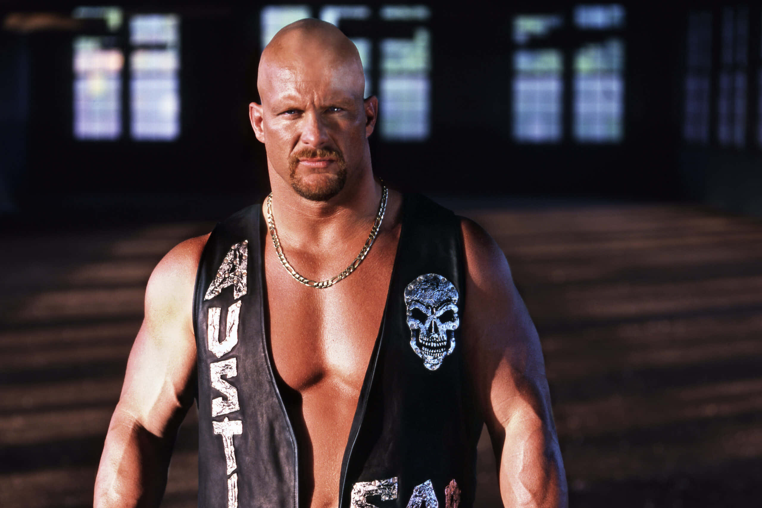 WWE 桌布 壁紙 — 一個光頭肌肉男，留著山羊鬍，穿著帶有骷髏頭和文字補丁的黑色背心，戴著鏈條項鍊，在一個光線昏暗、有窗戶的房間裡。