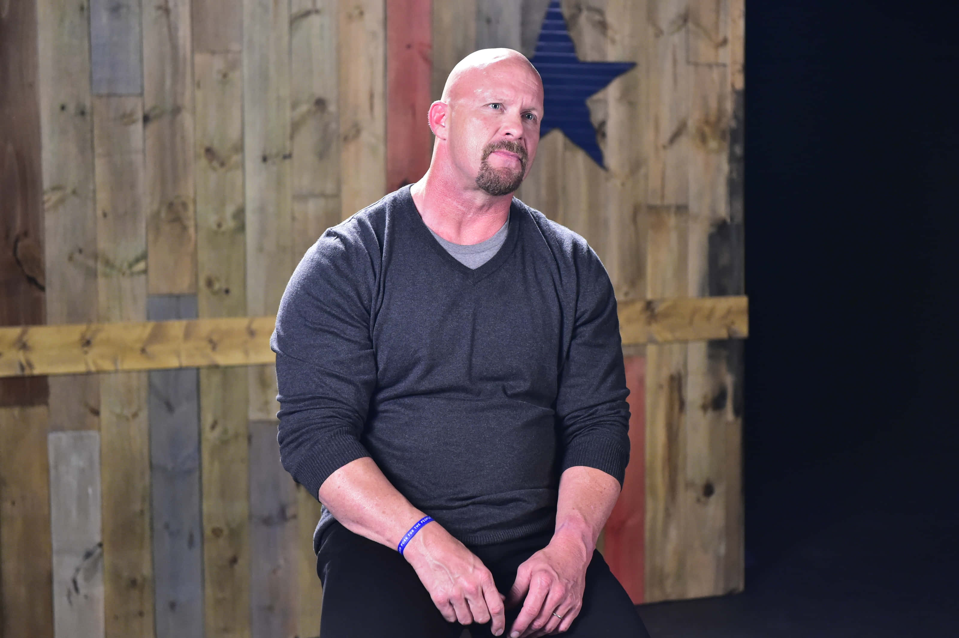 WWE 桌布 壁紙 — 一個光頭肌肉男留著山羊鬍，平靜地坐在一面有藍色星星的質樸木牆前。