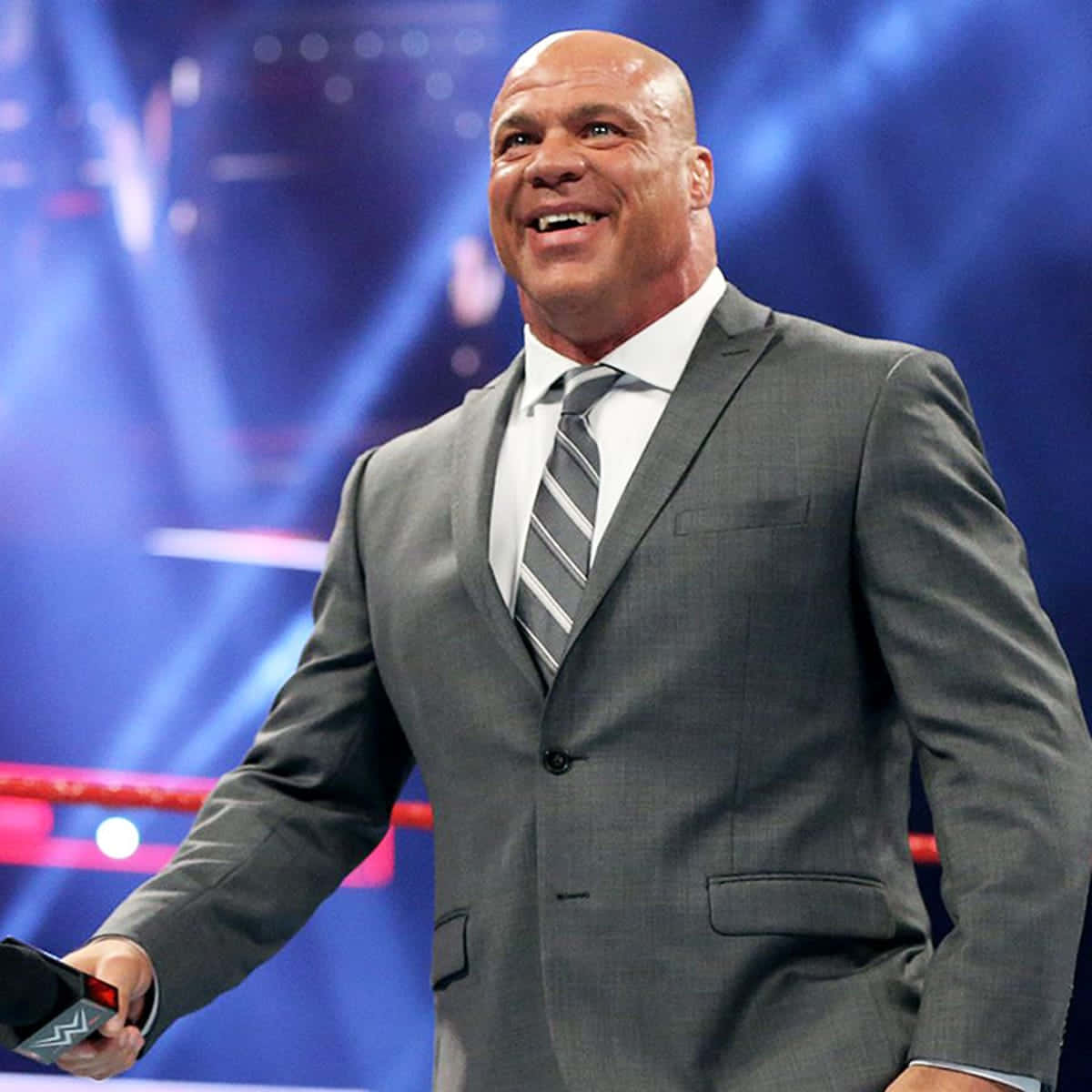 WWE 桌布 壁紙 — 一個光頭肌肉男穿著灰色西裝和條紋領帶，在明亮的藍色舞台燈光下，於摔角擂台內微笑著手持WWE麥克風。