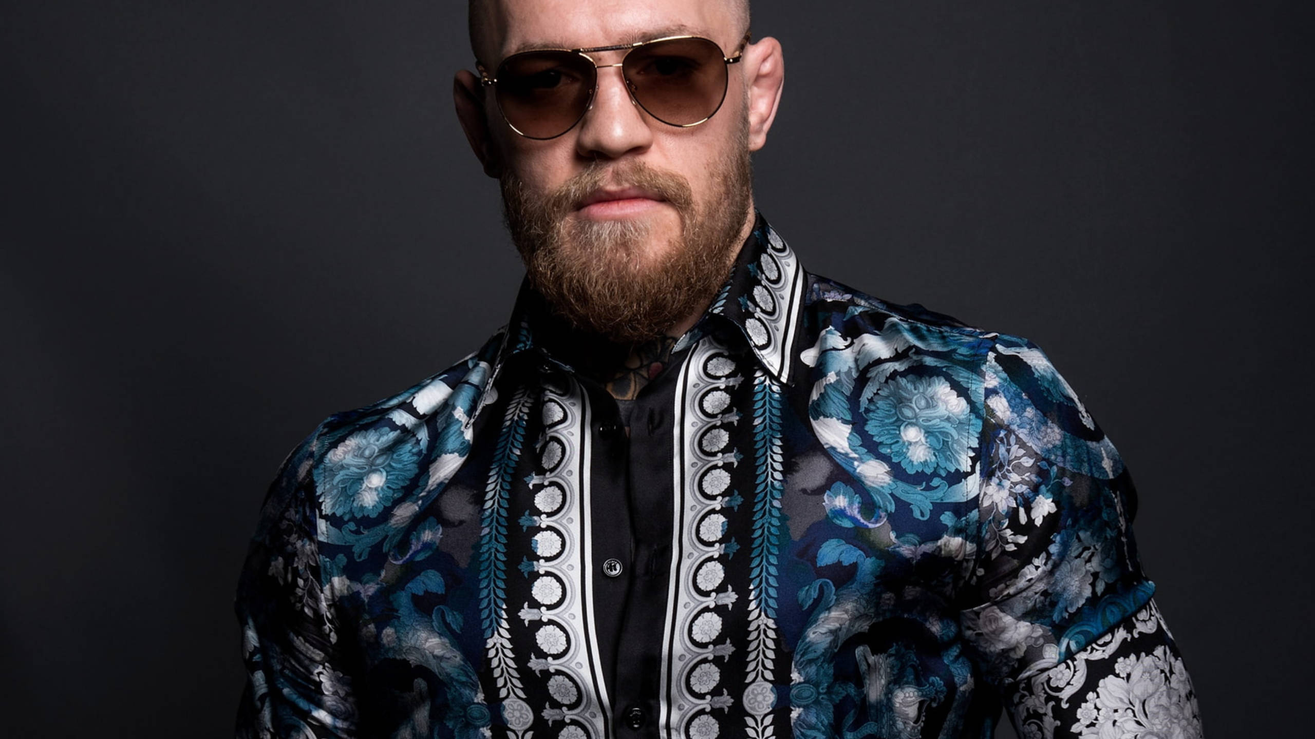 Mcgregor 桌布 壁紙 — 一個光頭鬍鬚男戴著飛行員太陽眼鏡，身穿大膽的藍色花卉圖案襯衫，背景深色。