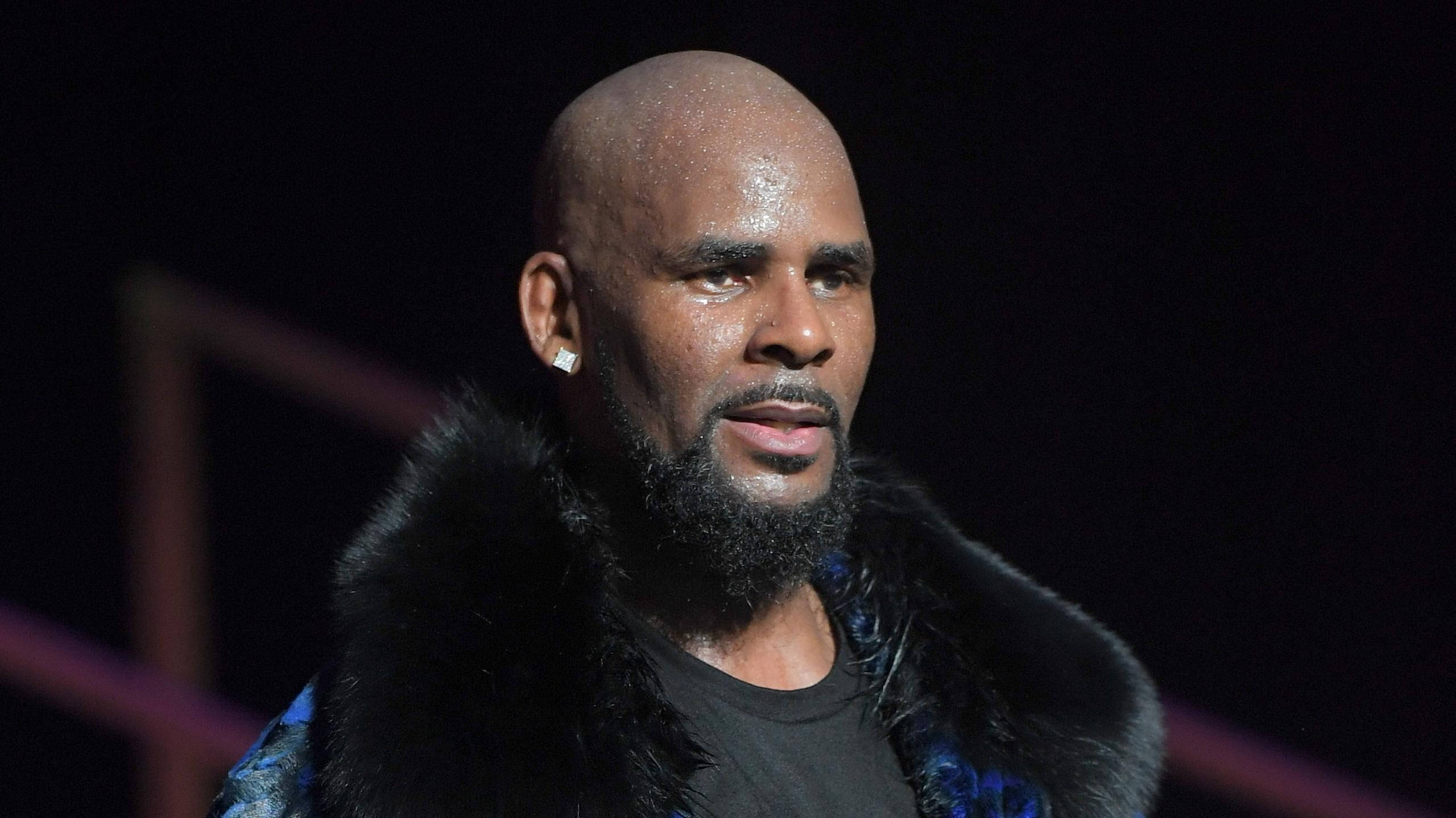 r kelly 桌布 壁紙 — 一個光頭鬍鬚男穿著毛領，站在舞台燈光下，背景黑暗。