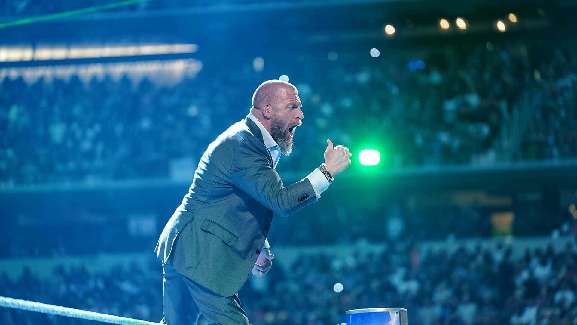WWE 桌布 壁紙 — 一個光頭鬍鬚男穿著西裝，在明亮的藍色舞台燈光下，在摔角場內激情咆哮，背景是模糊的人群。