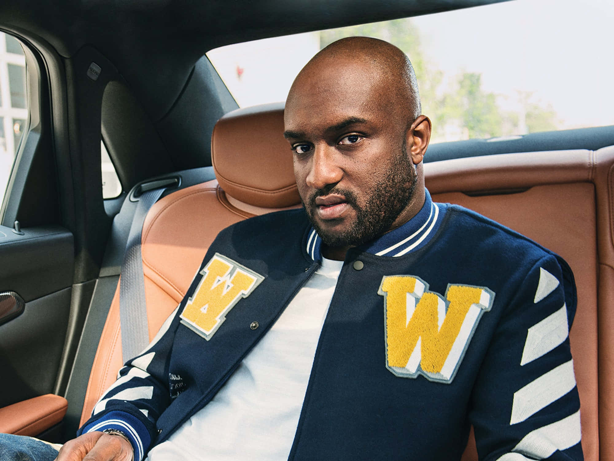 off white 桌布 壁紙 — 一個光頭鬍鬚男自信地坐在豪華車內，身穿海軍藍Varsity外套，上有黃色'W'字樣。