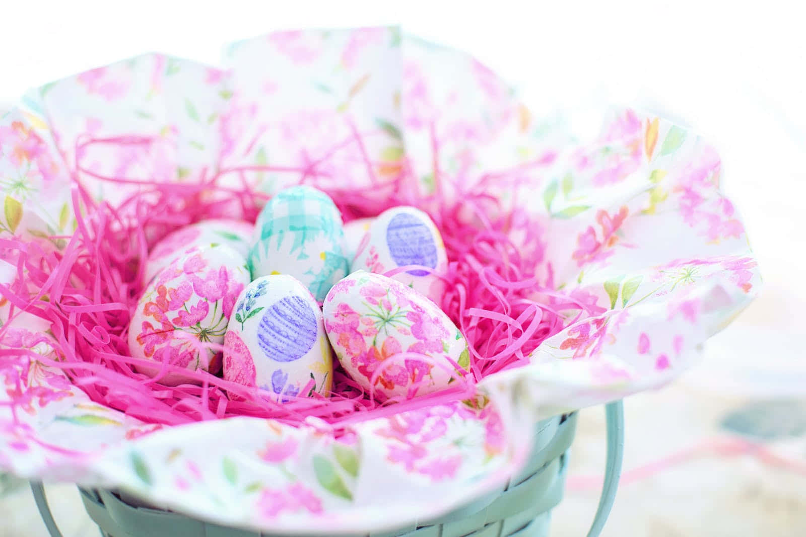 Pastel Easter 桌布 壁紙 — 一個內襯花卉圖案紙的籃子，裝著色彩繽紛、帶有圖案的復活節彩蛋，安放在亮粉紅色的碎紙中。