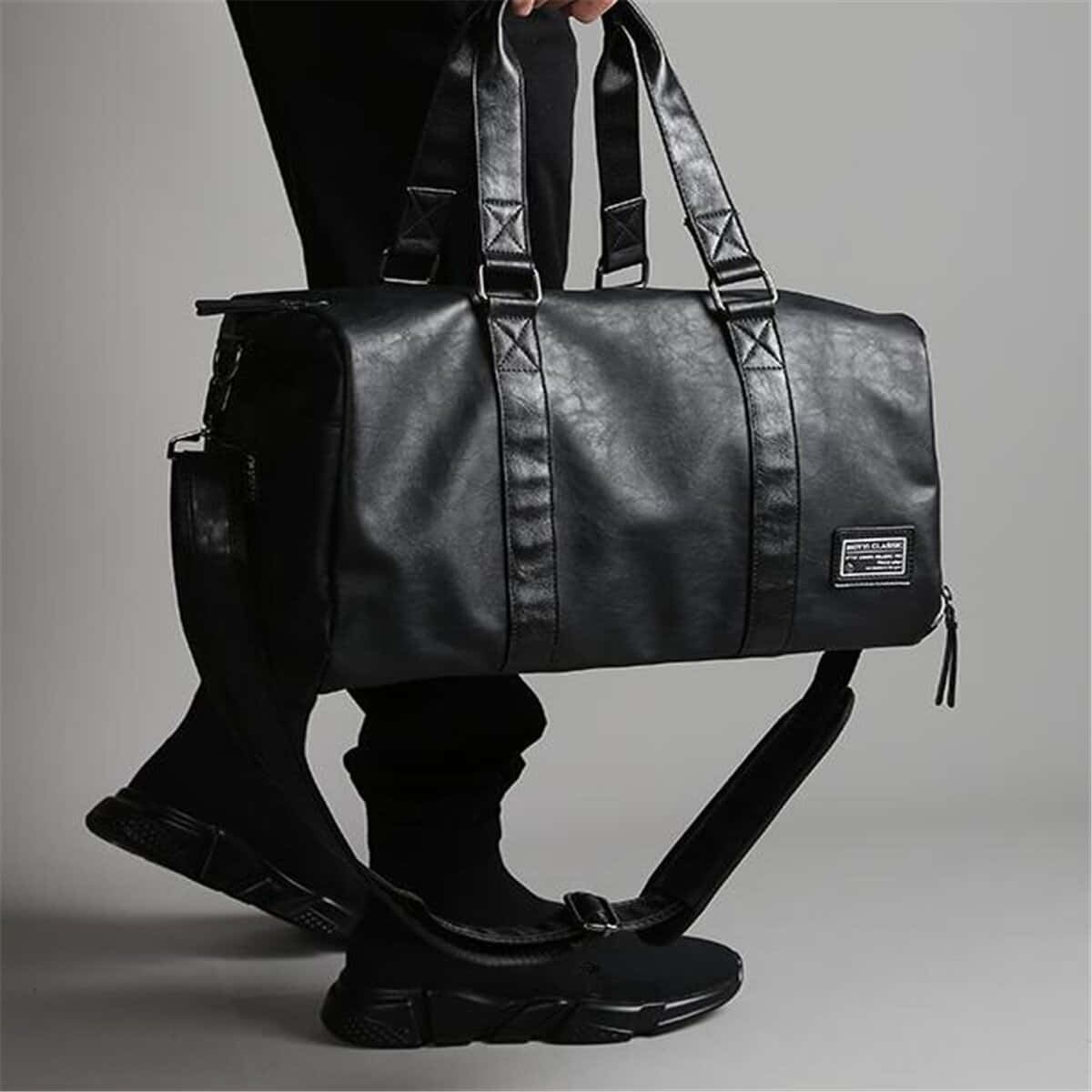Black Leather 桌布 壁紙 — 一個全身黑色的人，在灰色中性背景前，拿著一個帶有肩帶和拉鍊細節的時尚黑色皮革行李袋。