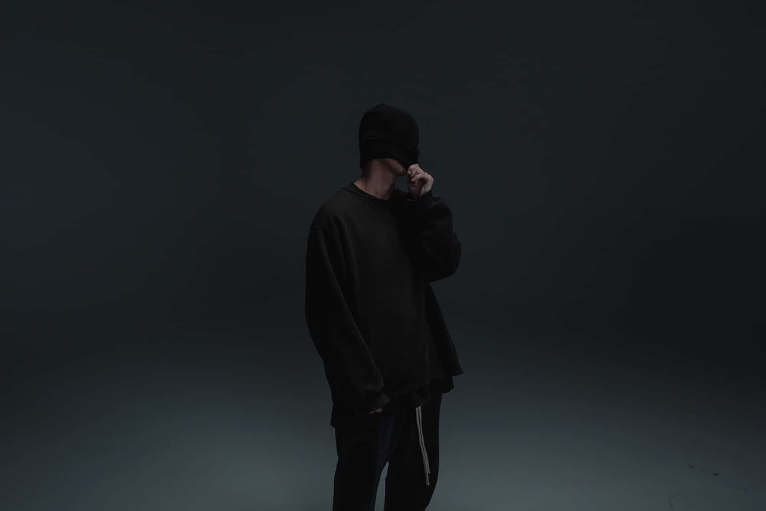 Nf Rapper 桌布 壁紙 — 一個全身黑色的人站在昏暗的攝影棚中，手靠近臉部，背景為深色漸變。
