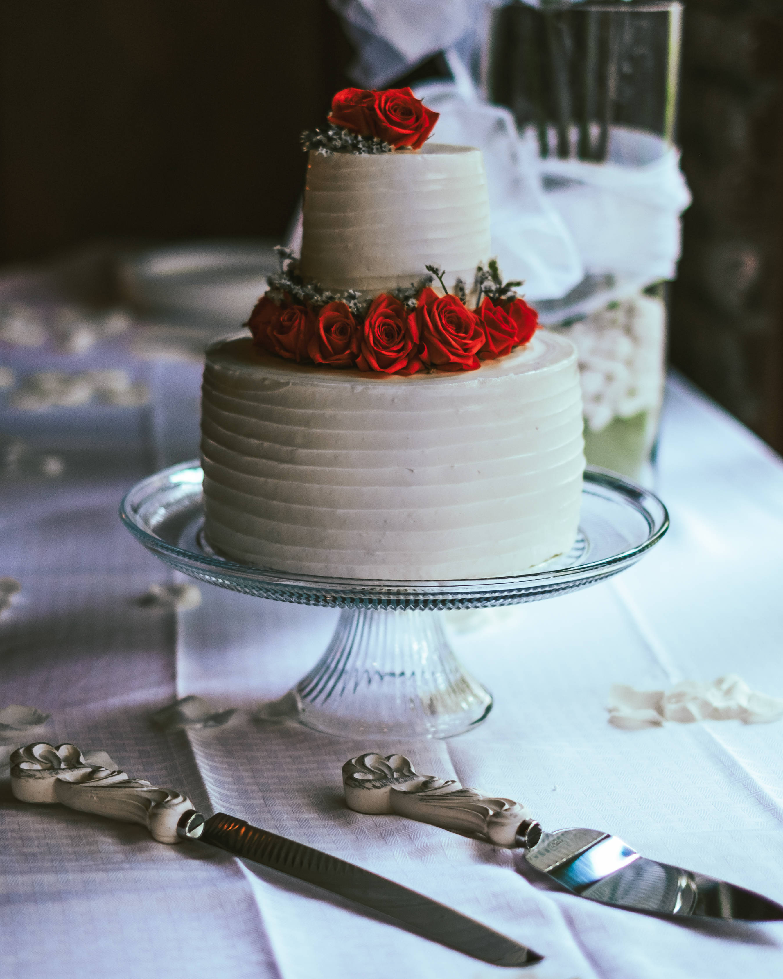 Wedding 桌布 壁紙 — 一個兩層白色婚禮蛋糕，飾以鮮豔的紅玫瑰，優雅地放在玻璃架上，置於白色桌布上，附近有復古蛋糕刀。