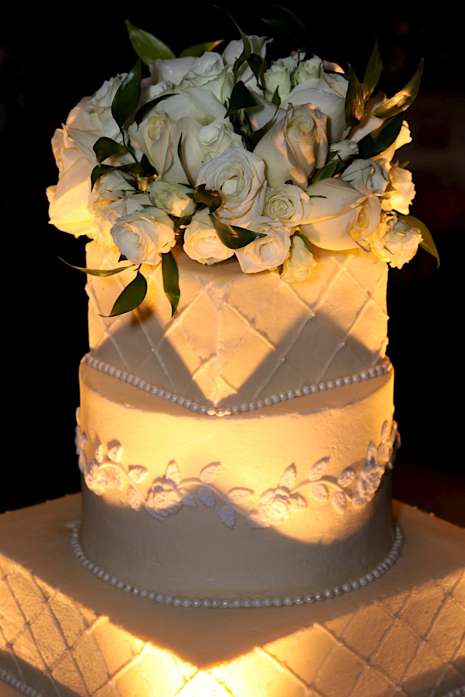 Wedding Cake 桌布 壁紙 — 一個兩層白色婚禮蛋糕，飾有玫瑰和珍珠狀珠子，在溫暖的金色燈光下，襯托著深色背景。