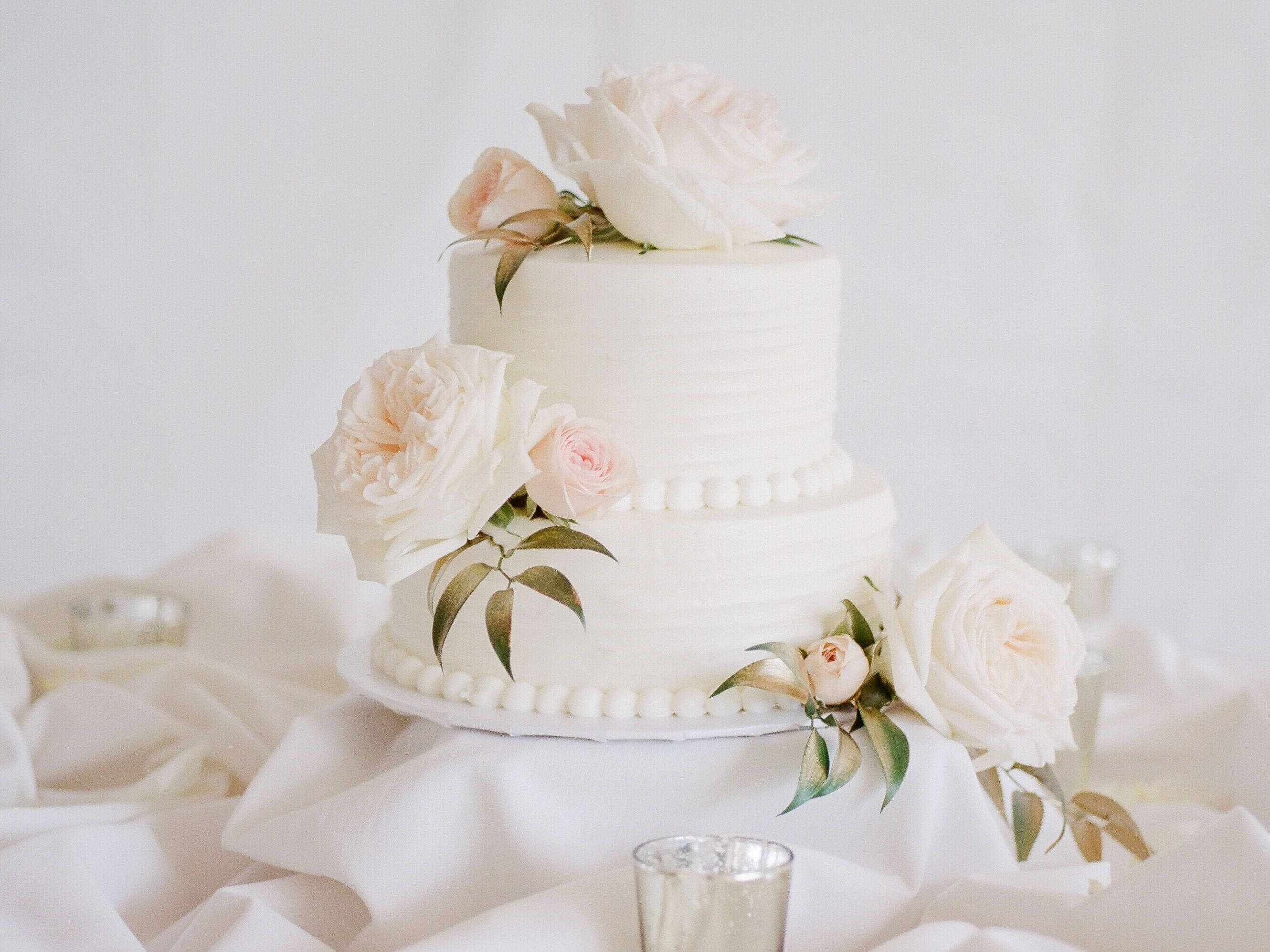 Wedding 桌布 壁紙 — 一個兩層白色結婚蛋糕，飾以柔和粉紅和象牙色玫瑰，優雅地放置在垂墜布料上。