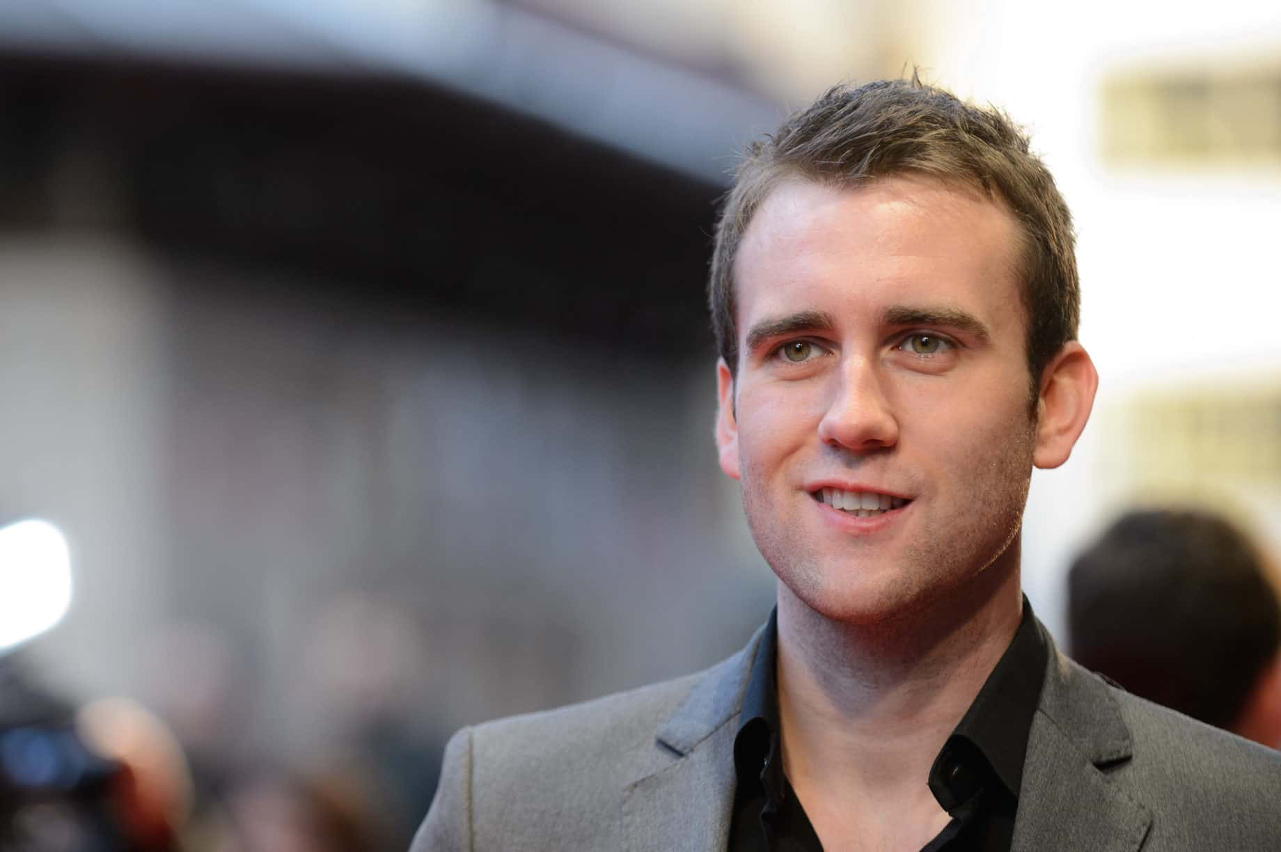 Neville Longbottom 桌布 壁紙 — 一個公開活動中，微笑男士穿著灰色西裝和黑色襯衫的 close-up 肖像，背景燈光模糊。