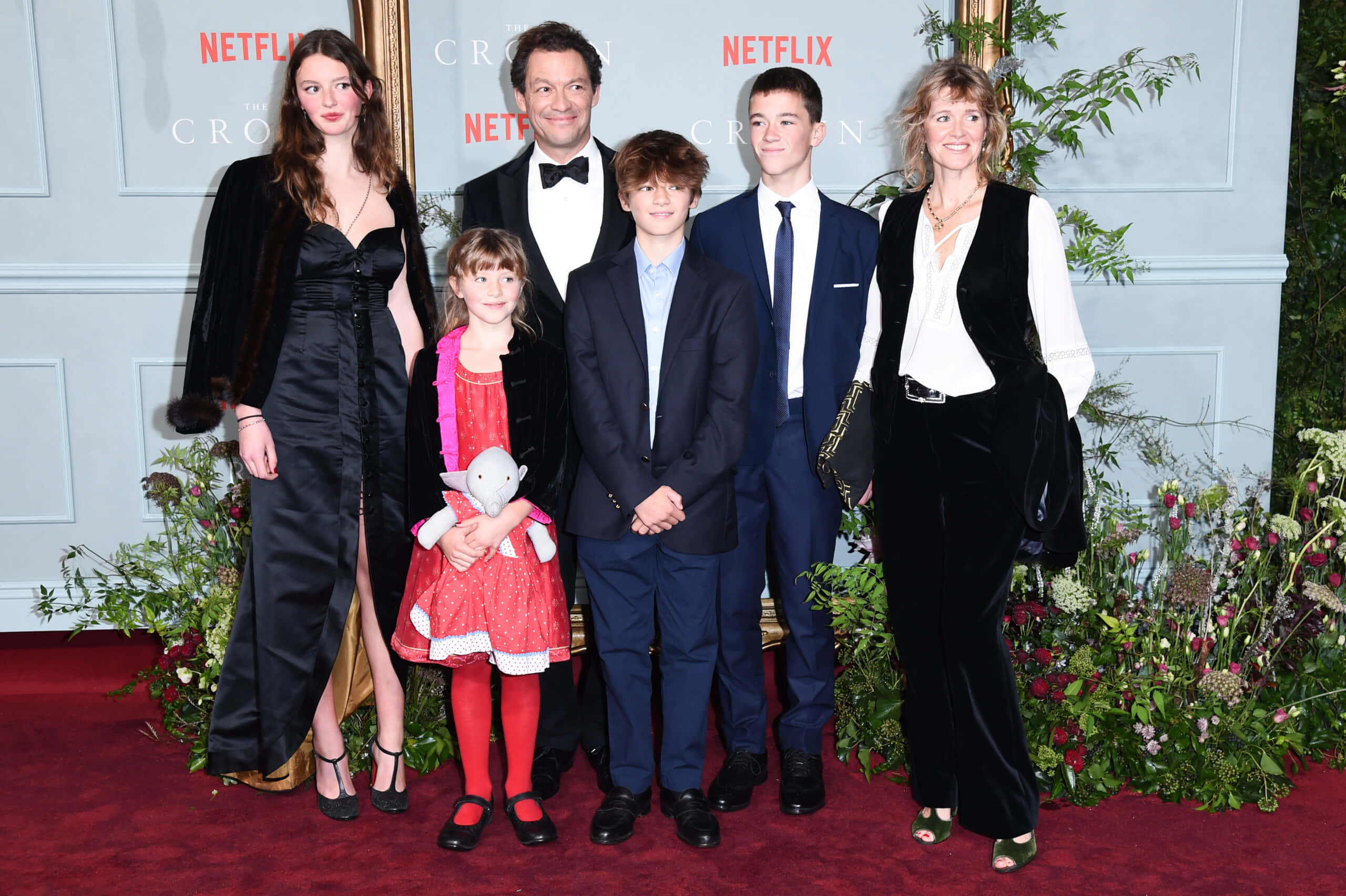 Dominic West 桌布 壁紙 — 一個六人家庭在品牌背景前，於兩旁擺放花卉佈置的紅地毯上正式擺姿勢。