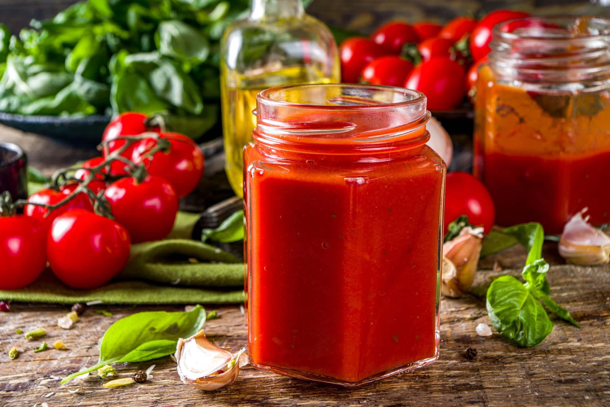 Red Sauce 桌布 壁紙 — 一個六角形玻璃瓶裝滿鮮紅色番茄醬，放在質樸的木桌上，周圍是新鮮羅勒、小番茄、大蒜和橄欖油。