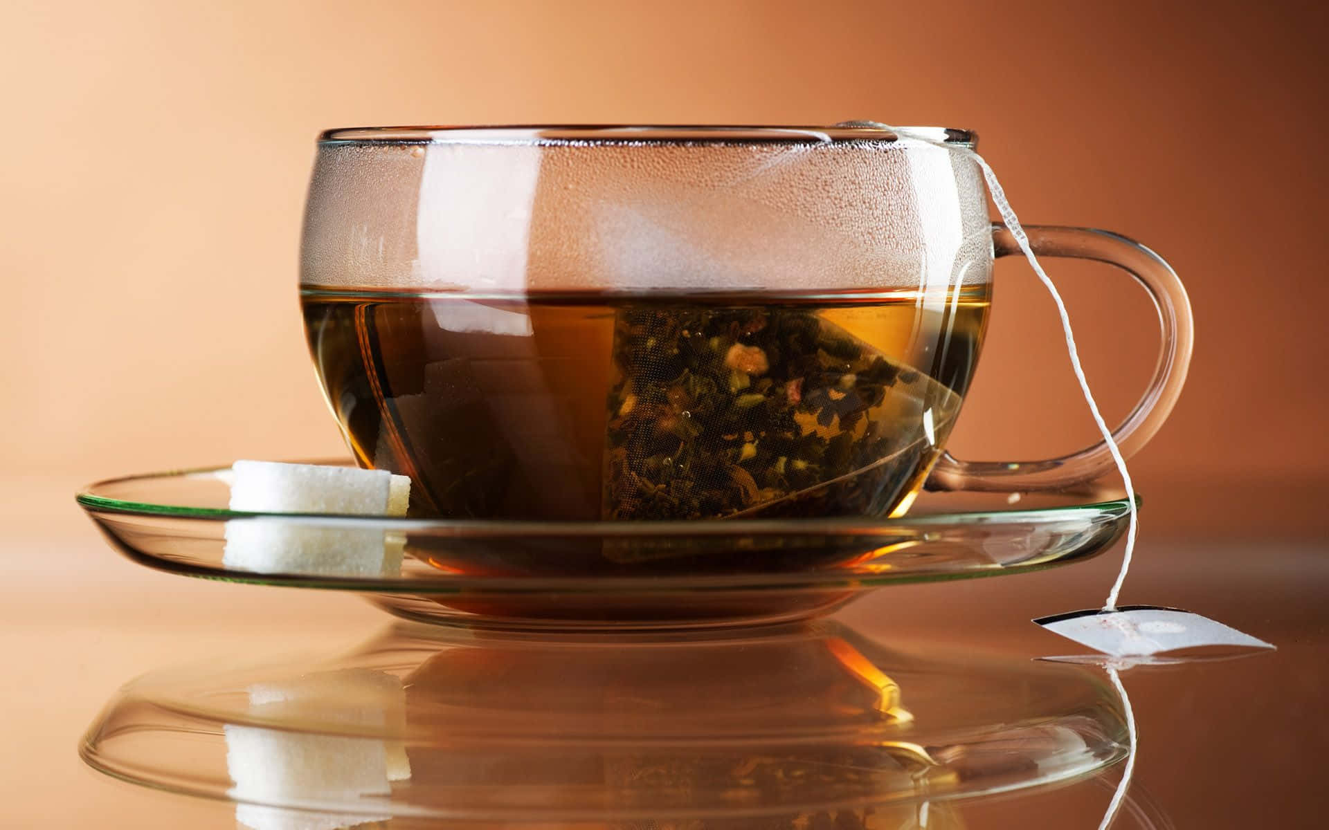 tea 桌布 壁紙 — 一個冒煙的玻璃茶杯，內有可見的茶包和方糖，放在一個反光的碟子上，背景是溫暖的漸變色。
