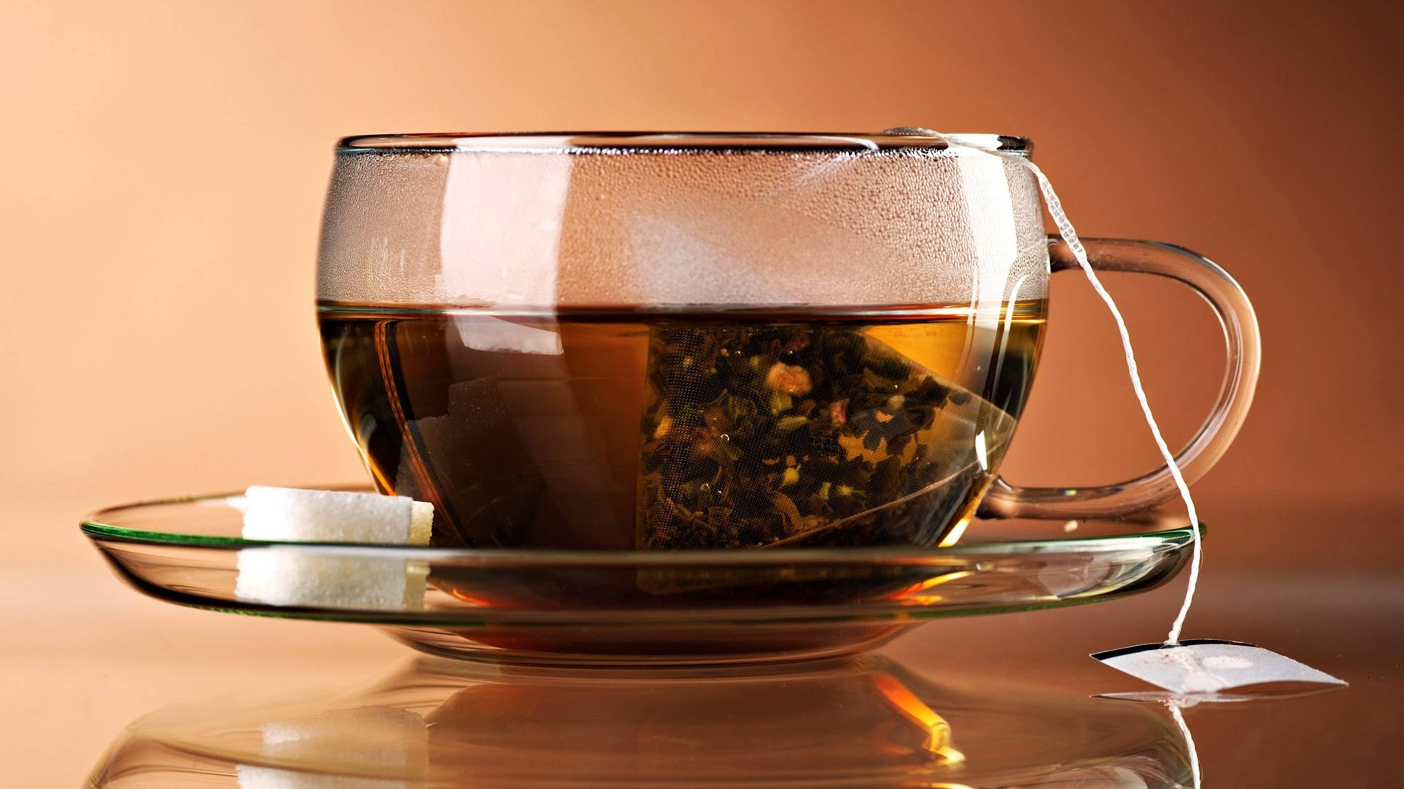 tea 桌布 壁紙 — 一個冒煙的玻璃茶杯，內有可見的茶包和方糖，放在相配的茶托上，背景是溫暖的棕色。