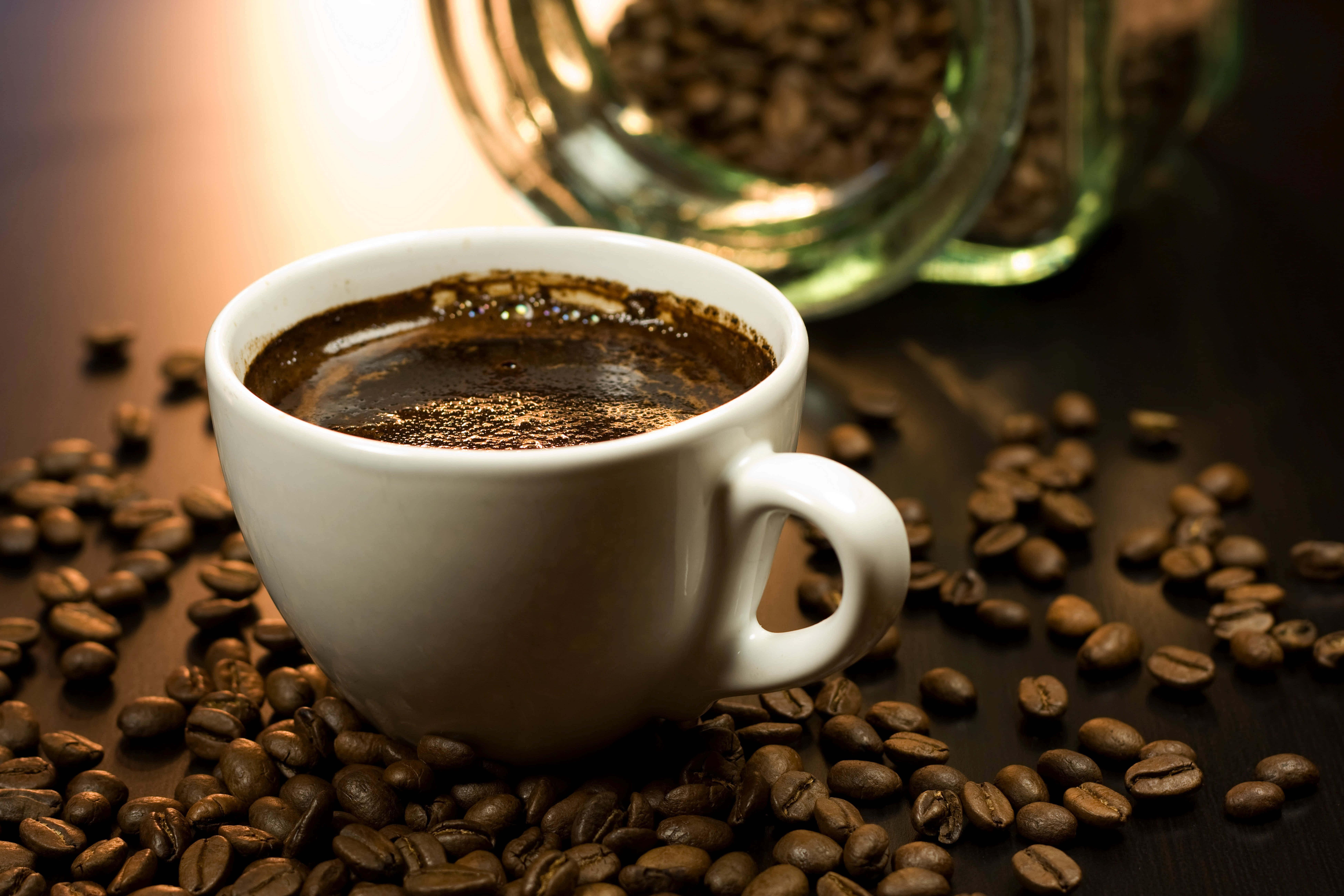 Black Coffee 桌布 壁紙 — 一個冒煙的白色咖啡杯，周圍是烘焙咖啡豆，放在深色木質表面上，旁邊有一個玻璃罐，在柔和溫暖的光線中。