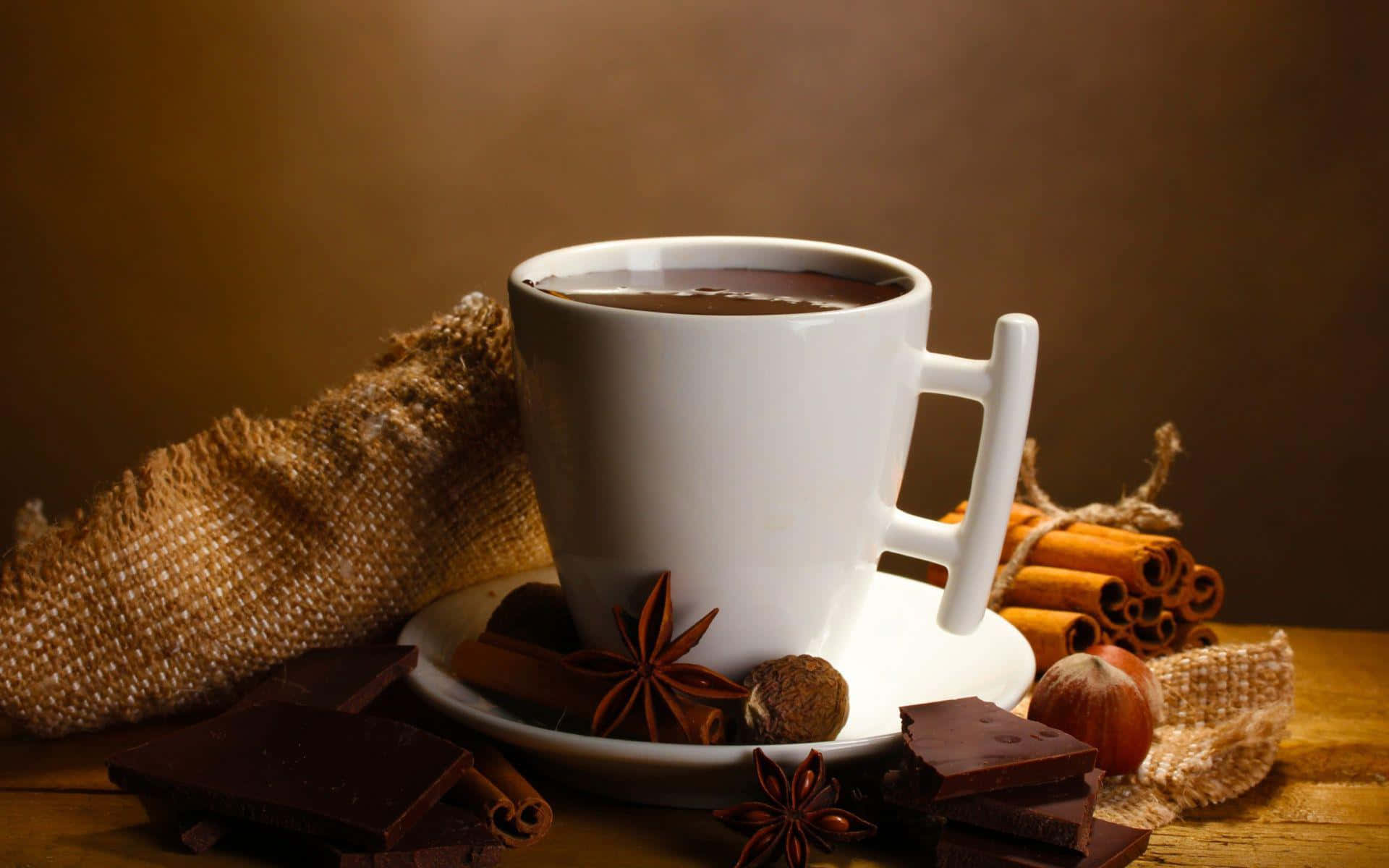 Brown Chocolate 桌布 壁紙 — 一個冒煙的白色馬克杯熱巧克力，周圍有巧克力塊、肉桂棒和八角，放在鄉村風格的木桌上。
