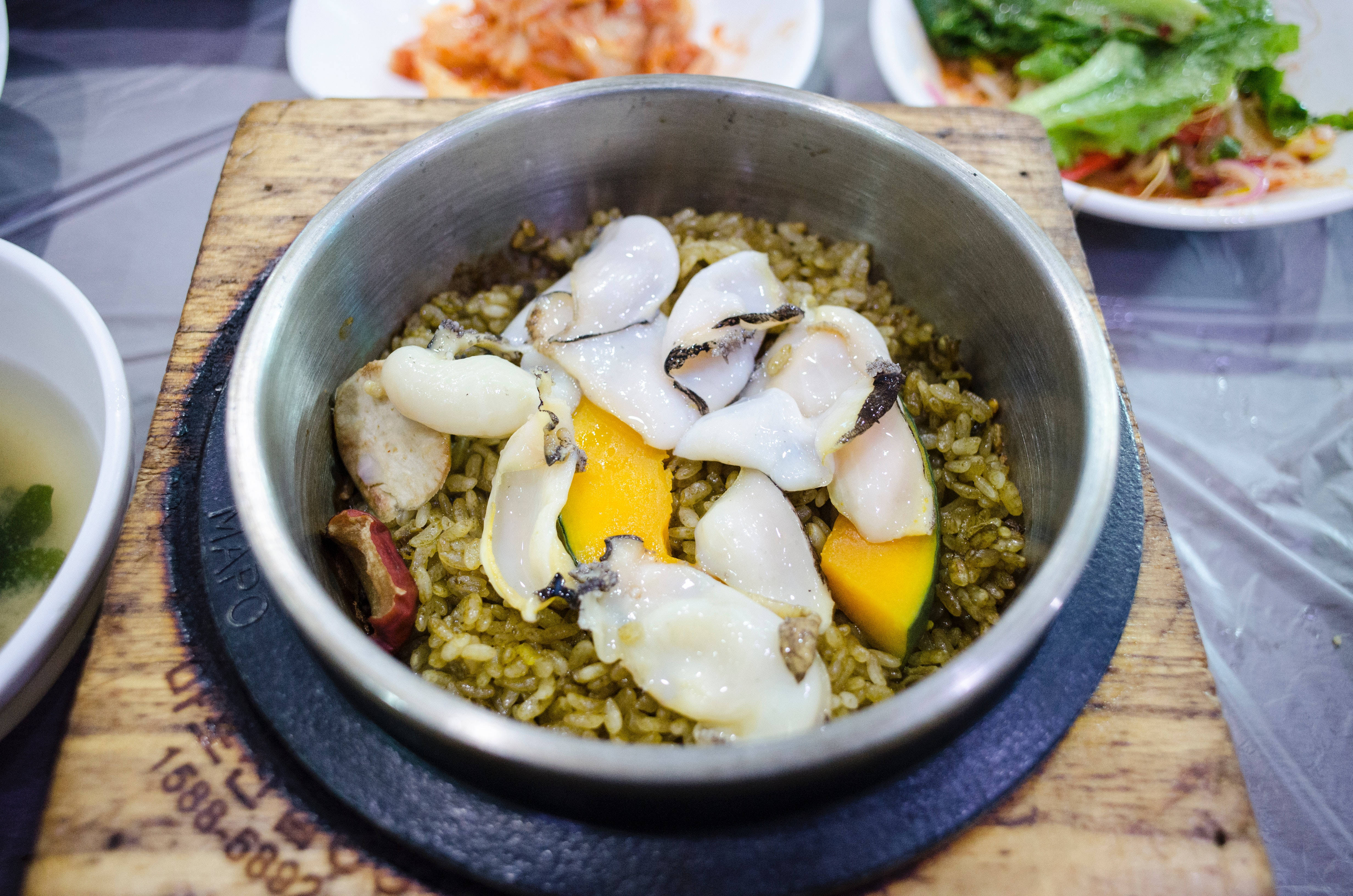 Rice 桌布 壁紙 — 一個冒煙的石鍋，盛載著韓國海鮮飯，配以生蠔、芒果和青菜，放在木板上。