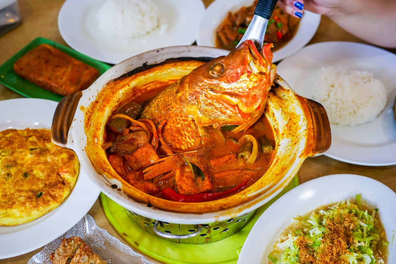 Food Photography 桌布 壁紙 — 一個冒煙的陶鍋，裡面裝著辛辣的魚咖哩，用鉗子夾起整條魚，周圍是米飯和配菜。