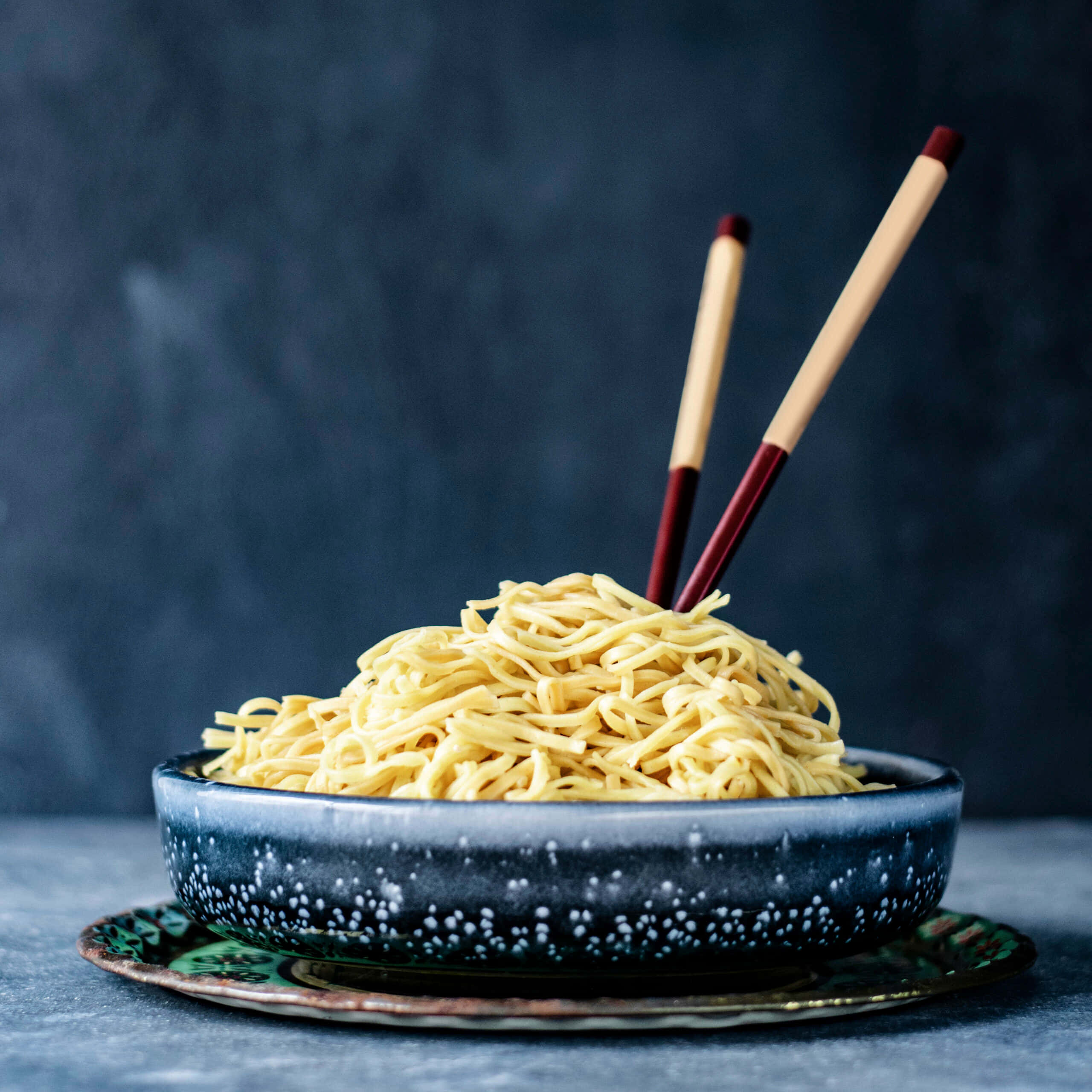 Noodles 桌布 壁紙 — 一個冒煙的黃色麵條碗，放在有斑點的深藍色陶瓷碗中，筷子擱在上面，背景是陰鬱的灰色。