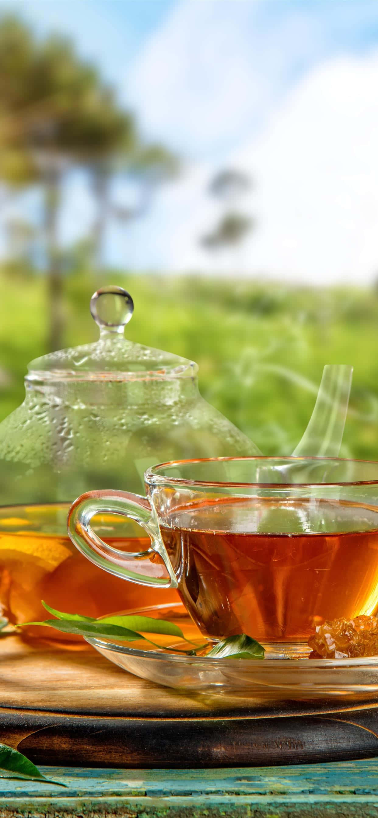 咖啡動圖 桌布 壁紙 — 一個冒著蒸氣的玻璃茶杯和茶壺放在戶外一個質樸的木托盤上，背景是明亮的藍天和柔焦的綠樹。
