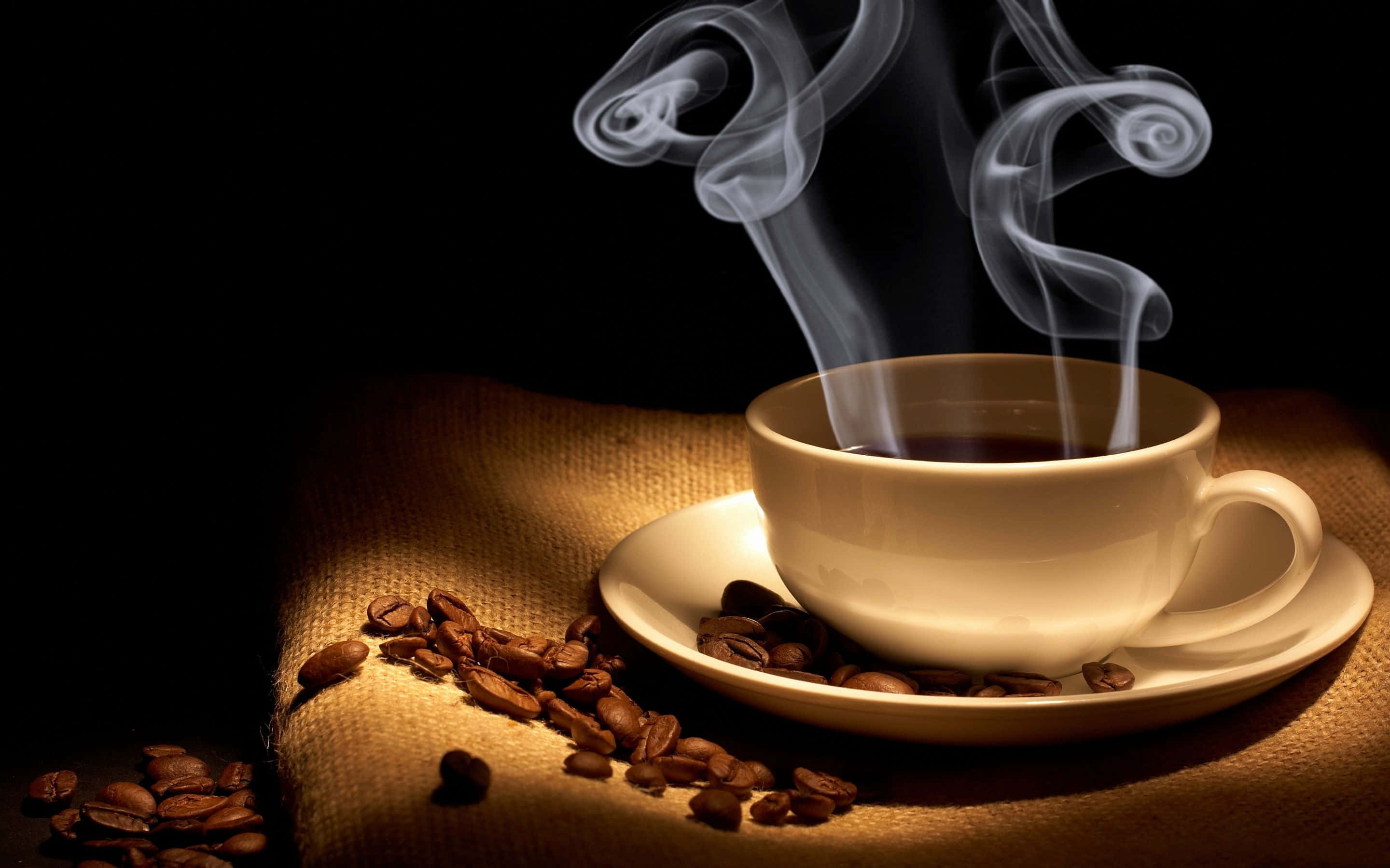 Coffee Aesthetic 桌布 壁紙 — 一個冒著蒸氣的白咖啡杯放在碟子上，碟子置於麻布上，周圍散落著咖啡豆，在黑色背景的暖光下閃閃發光。