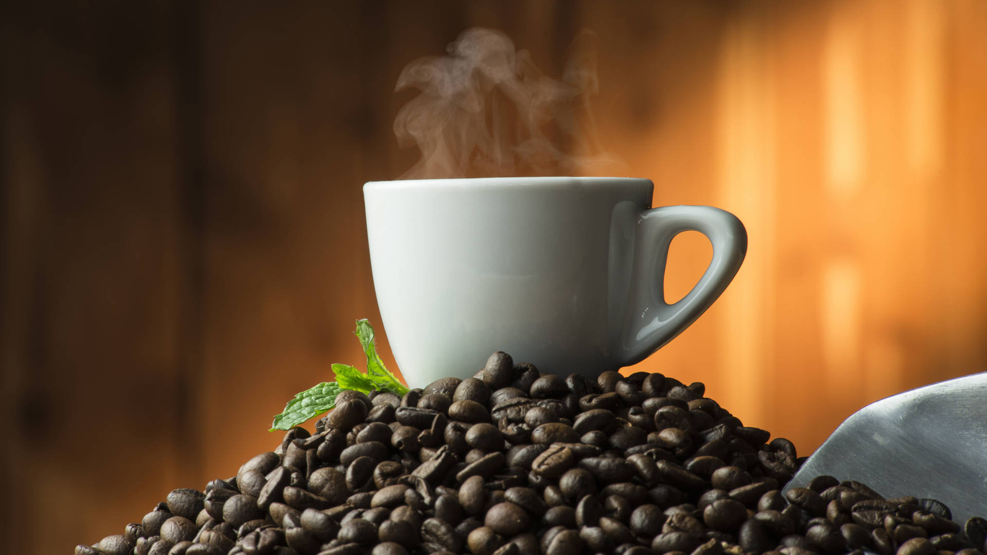 Coffee Aesthetic 桌布 壁紙 — 一個冒著蒸氣的白色咖啡杯放在一堆深烘焙咖啡豆上，旁邊有一片新鮮薄荷葉，背景是溫暖的木質。