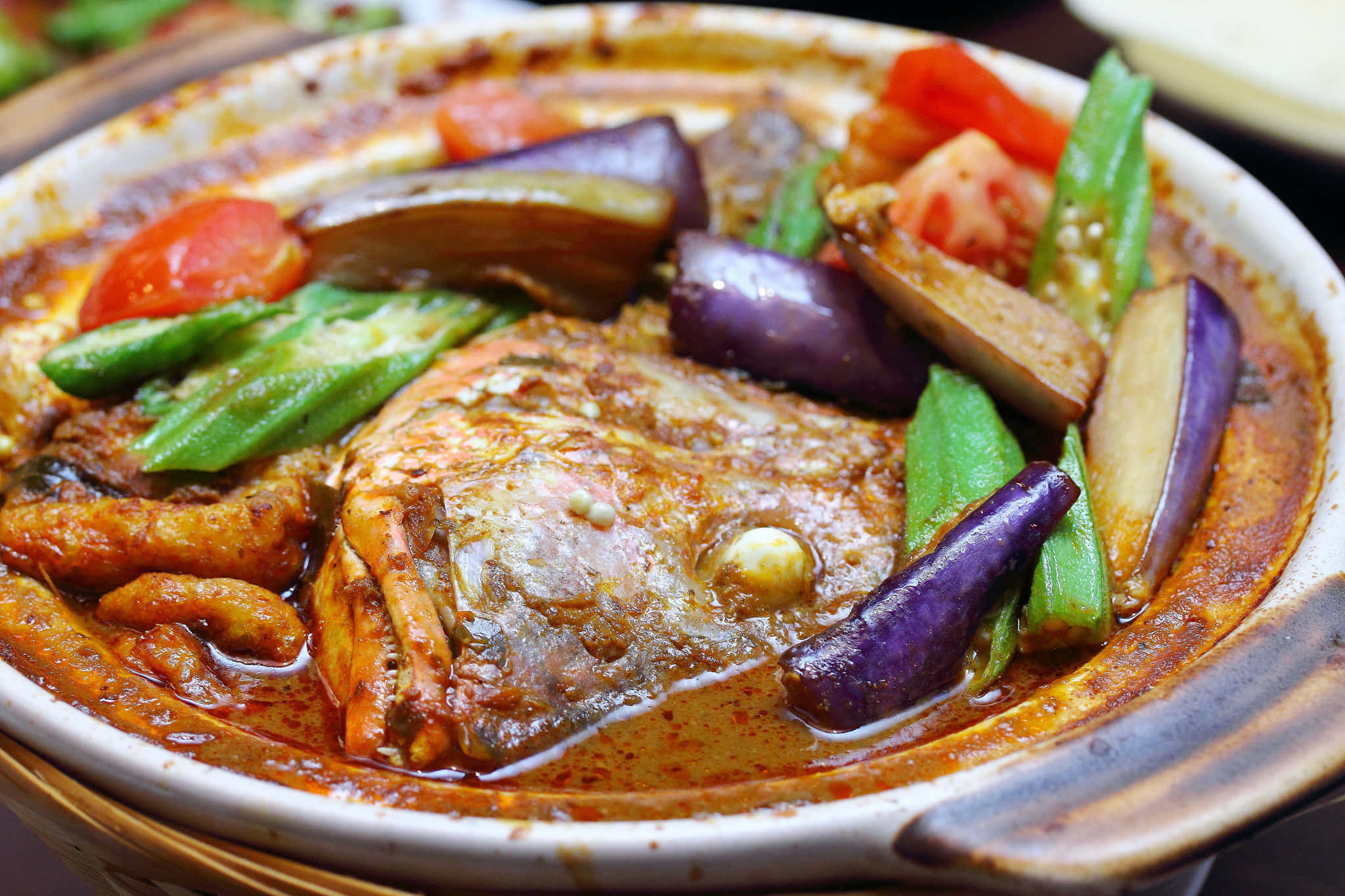 Food Photography 桌布 壁紙 — 一個冒著蒸氣的砂鍋，盛滿了香料豐富的魚頭咖哩，周圍環繞著鮮豔的茄子、番茄和秋葵。