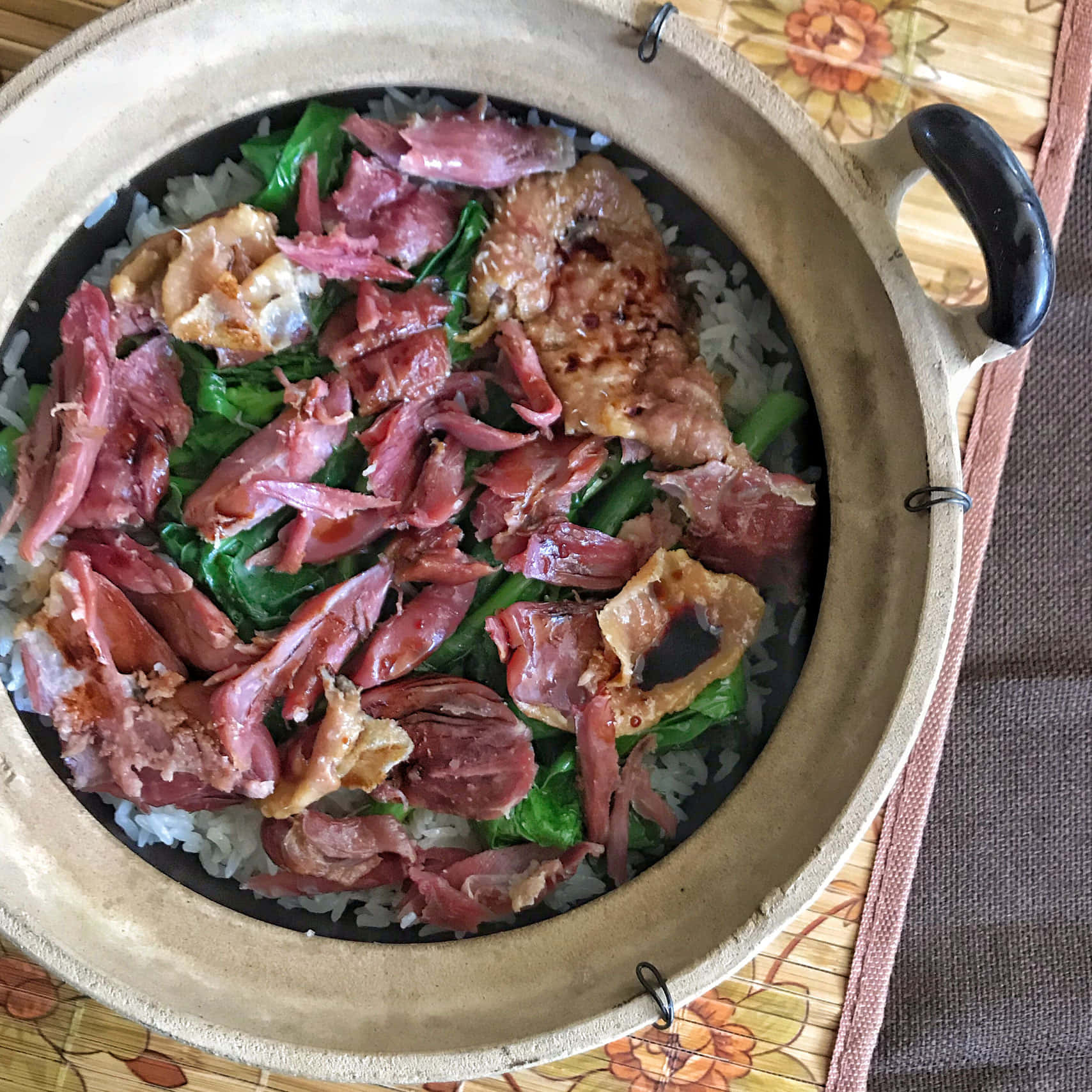 Food Photography 桌布 壁紙 — 一個冒著蒸氣的砂鍋，裡面裝滿了米飯，上面鋪著切片臘肉和綠色蔬菜，放在一個花卉圖案的餐墊上。