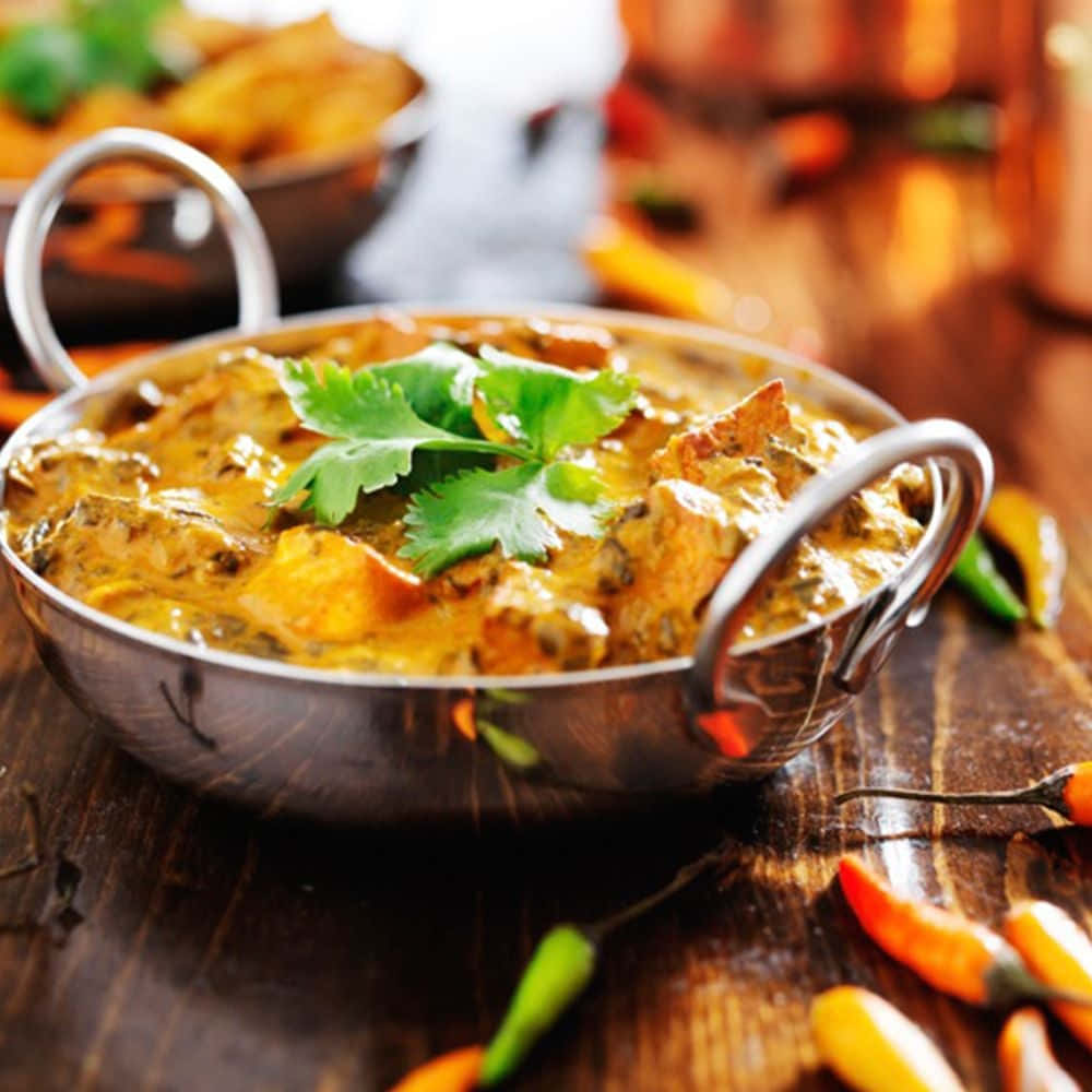 Indian Food 桌布 壁紙 — 一個冒著蒸氣的金屬碗，盛滿濃郁的黃咖哩雞和新鮮香菜，放在質樸的木桌上，周圍環繞著色彩繽紛的辣椒。