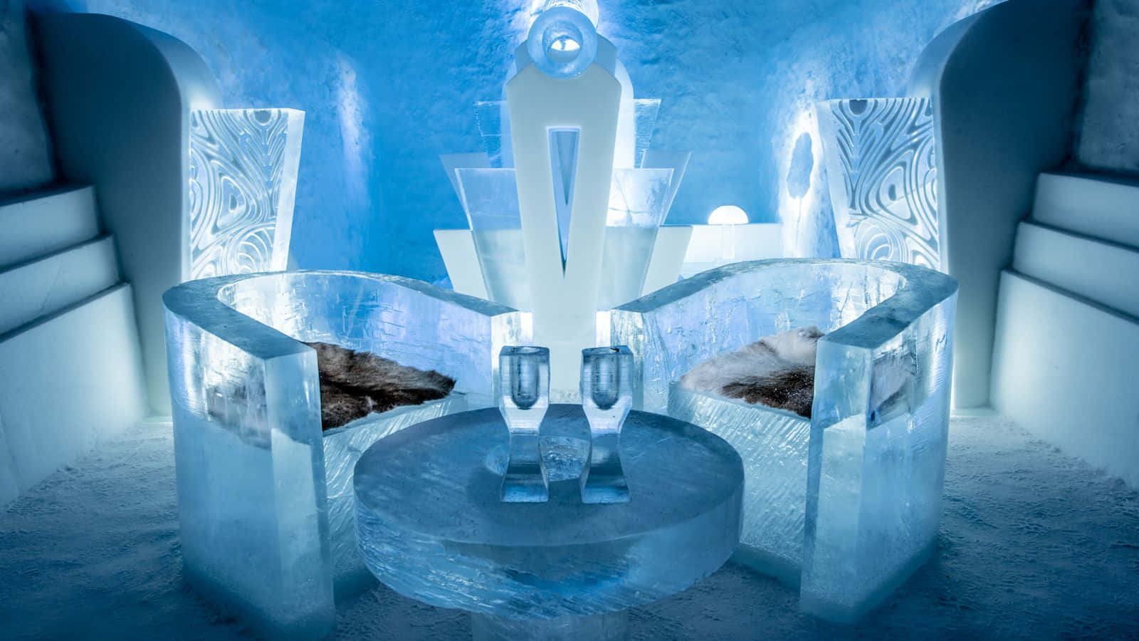 Ice Hotel 桌布 壁紙 — 一個冰封的王座廳，內有雕刻的冰椅和桌子，在冰洞中藍色燈光下閃耀。