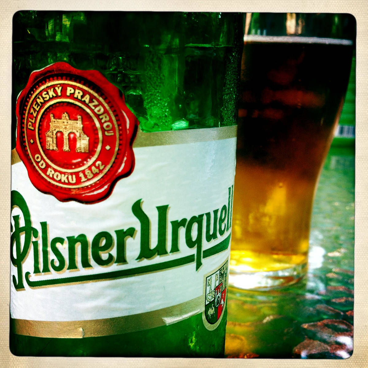 bottle 桌布 壁紙 — 一個冰涼的綠色 Pilsner Urquell 啤酒瓶，帶有紅色封條，旁邊放著一杯琥珀色啤酒，放在有紋理的吧檯面上。