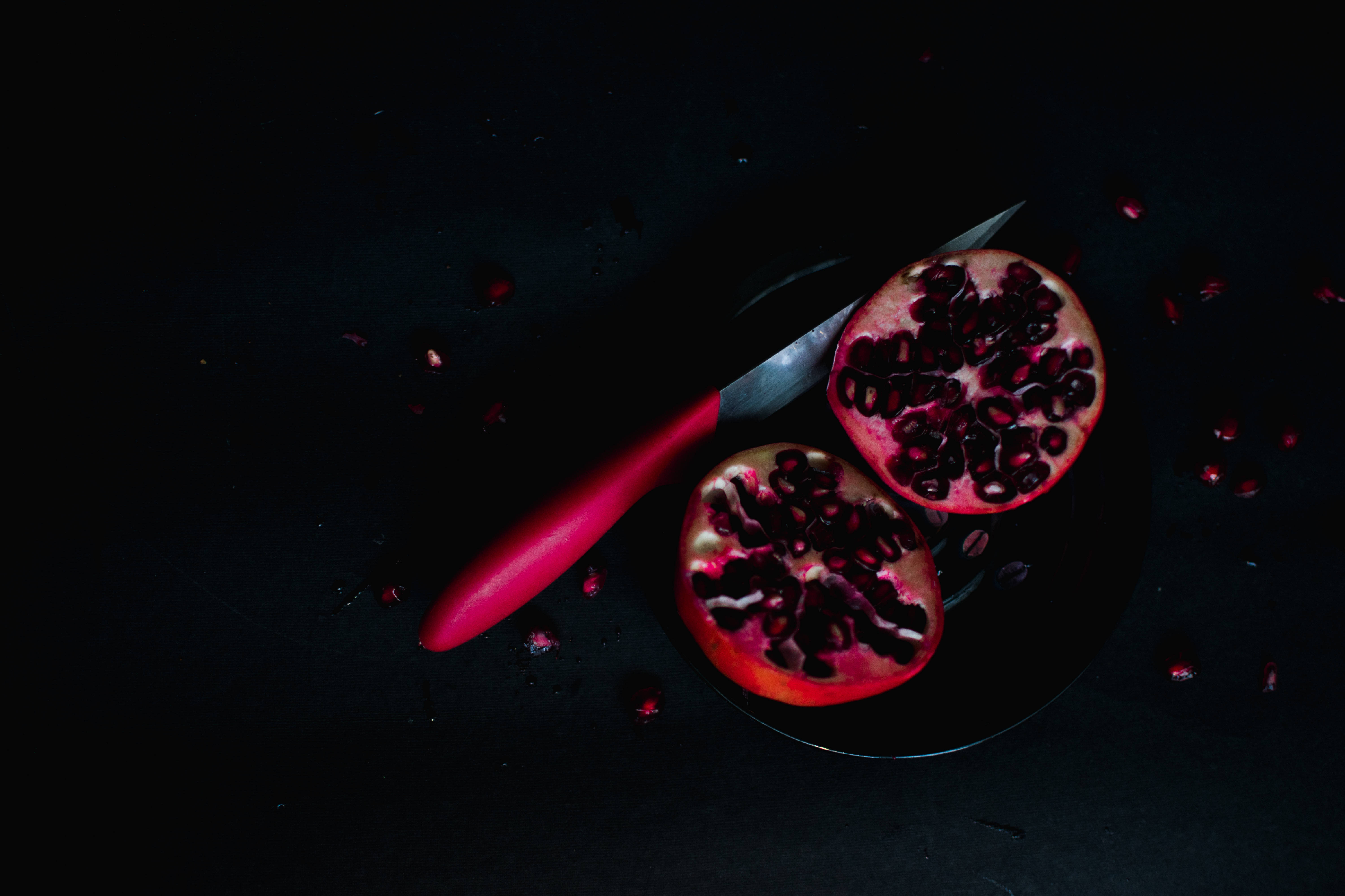 Fruits 桌布 壁紙 — 一個切開的石榴，配上一把紅柄刀，放在黑色表面上，種子在戲劇性的低光下散落。
