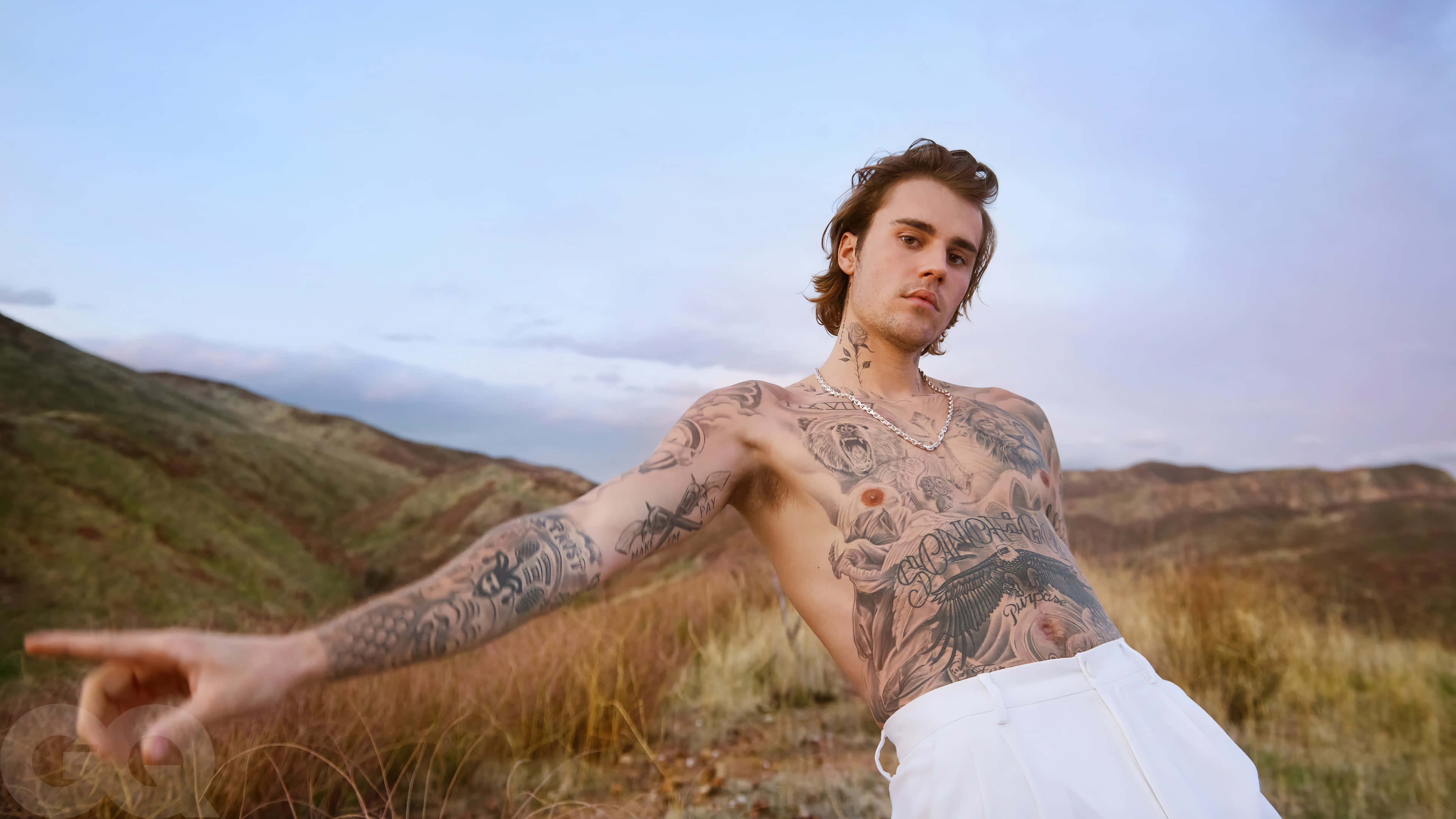 Justin Bieber 2021 桌布 壁紙 — 一個刺青男子穿著白色褲子，在黃昏時分對著柔和粉彩天空的丘陵景觀向前指。