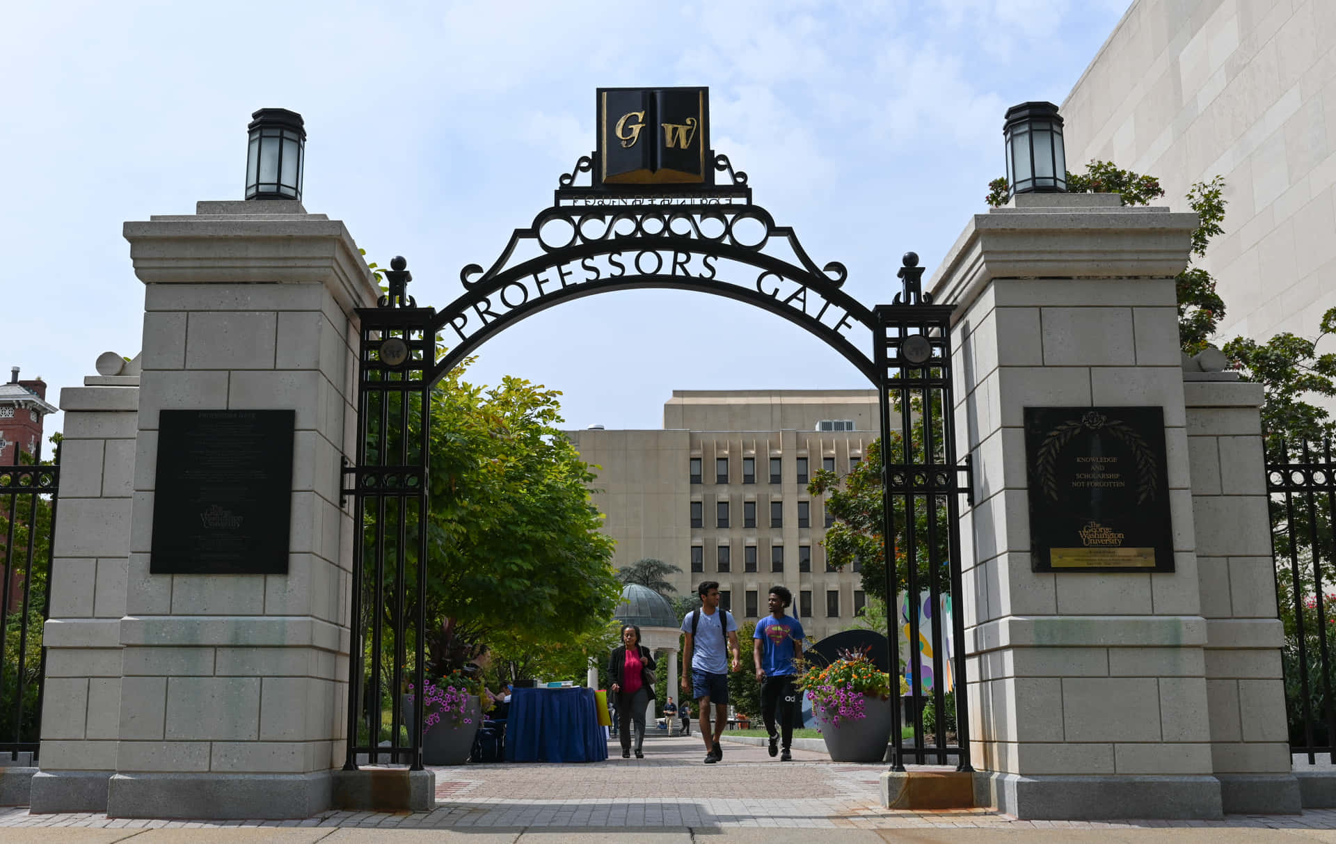  壁紙 — 一個刻有「PROFESSORS GATE」的大型石鐵拱門，框住行人走向大學建築物，背景是明亮的藍天。