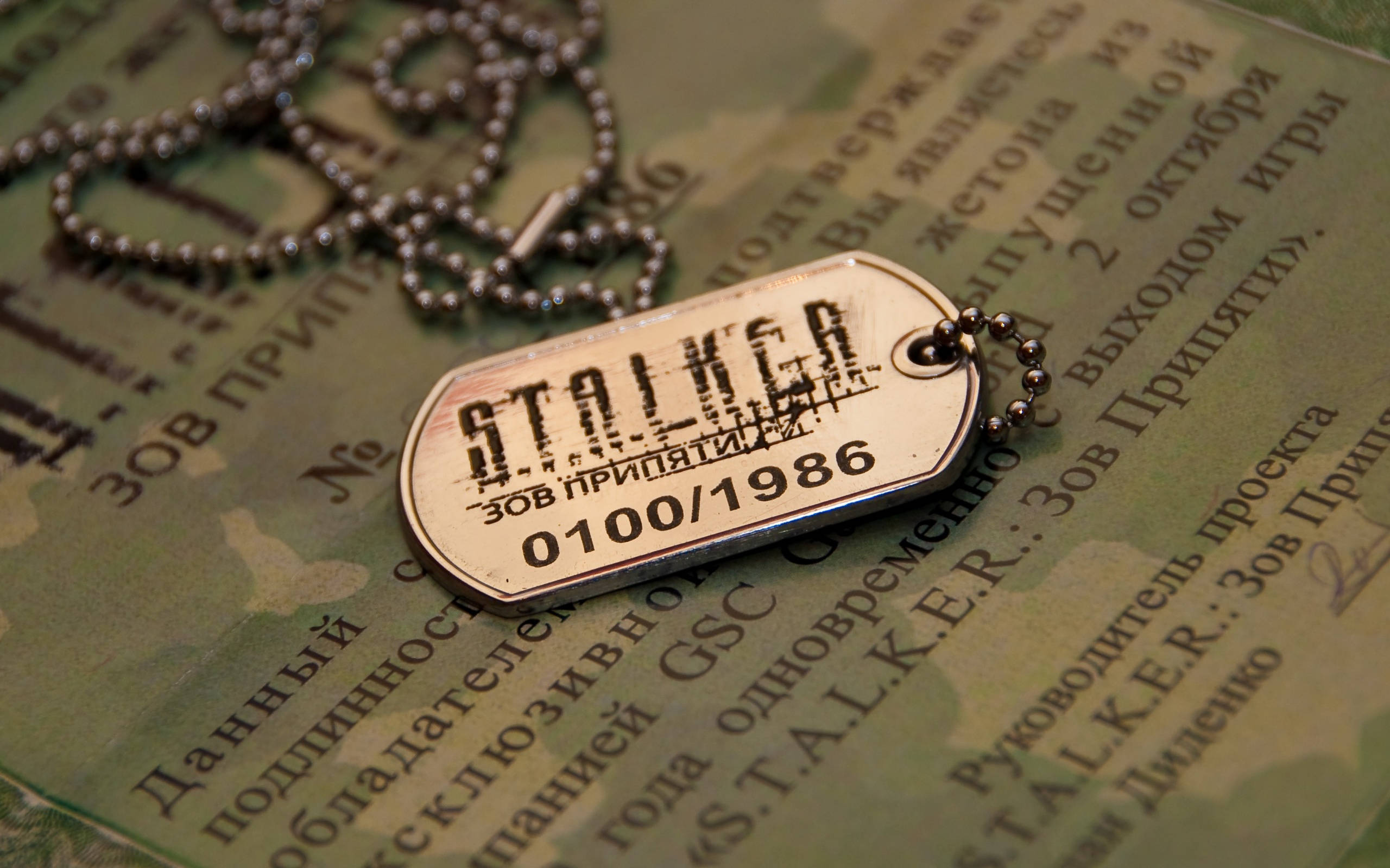 Stalker 桌布 壁紙 — 一個刻有「STALKER ЗОВ ПРИПЯТИ 0100/1986」的狗牌放在帶有西里爾文字的迷彩文件上。