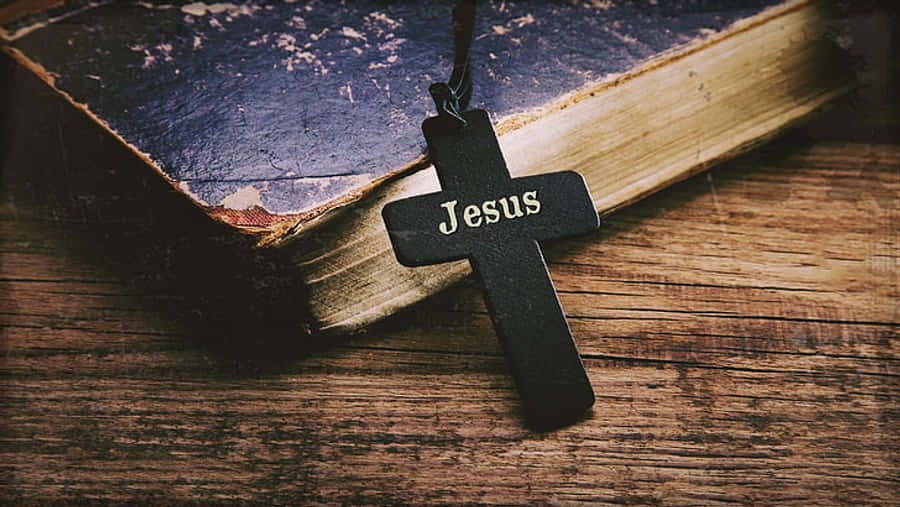 Jesus Name 桌布 壁紙 — 一個刻有『耶穌』的黑色十字架，安放在一張質樸木質表面上的破舊皮面聖經上。