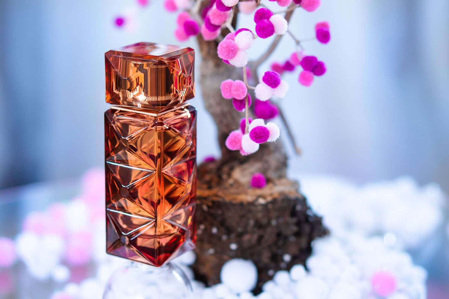 Spring Perfume 桌布 壁紙 — 一個刻面琥珀色香水瓶在雪白底座上，旁邊放著一棵夢幻的粉白色絨球樹，閃閃發光。