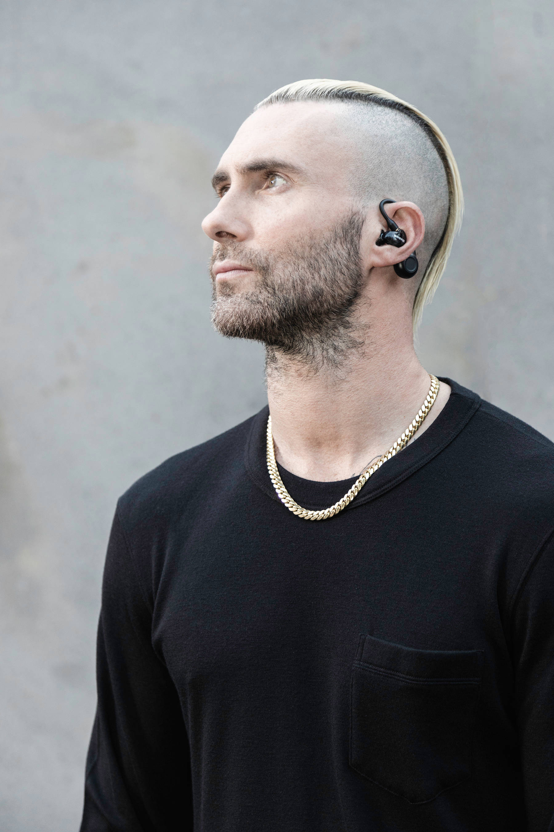 Adam Levine 桌布 壁紙 — 一個剃光頭、留著金色莫霍克髮型的男人，戴著黑色耳機和金鏈子，背景是灰色混凝土牆。