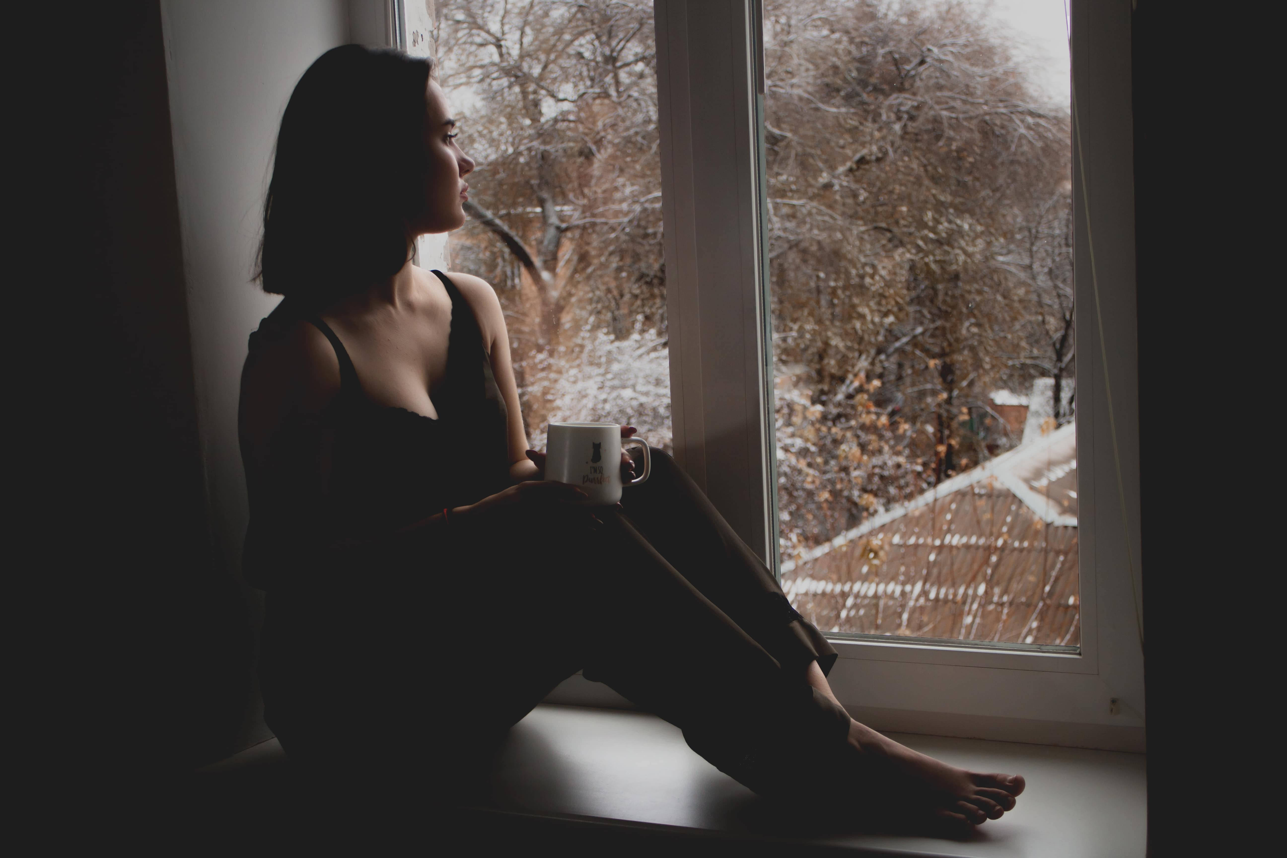 Sitting Alone 桌布 壁紙 — 一個剪影中的女人赤腳坐在窗台上，拿著杯子，凝視著外面白雪皚皚的冬日景色，樹木光禿禿的。