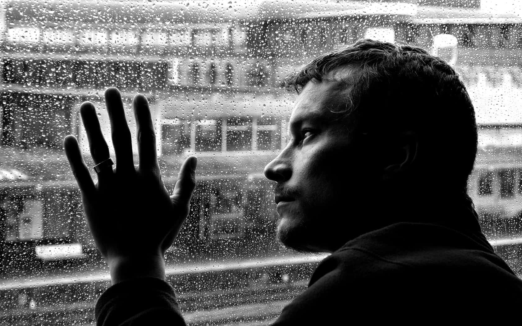 Sad Face 桌布 壁紙 — 一個剪影中的男人透過佈滿雨點的窗戶凝視著單色調模糊的城市景觀。