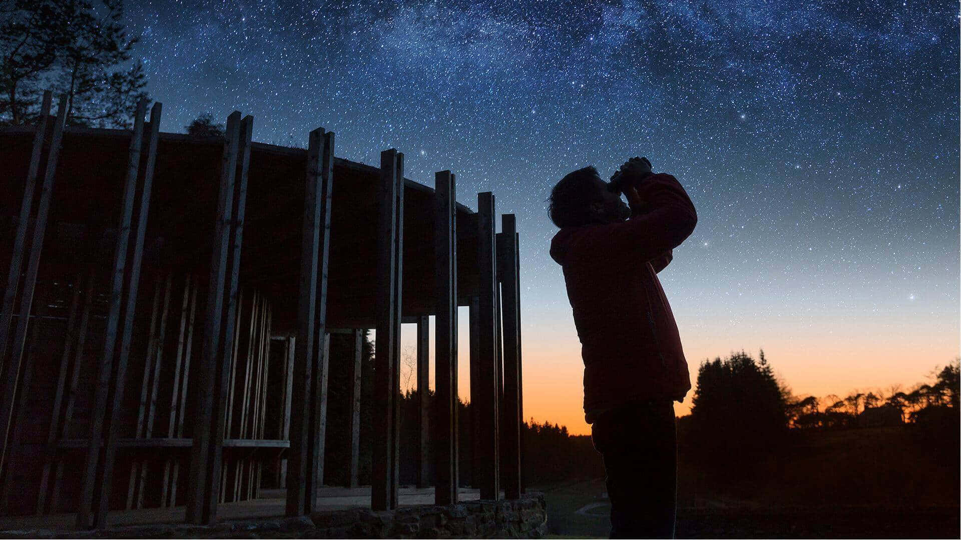 夜空 桌布 壁紙 — 一個剪影人物使用雙筒望遠鏡凝視木結構上方的銀河，暮色漸入夜晚。