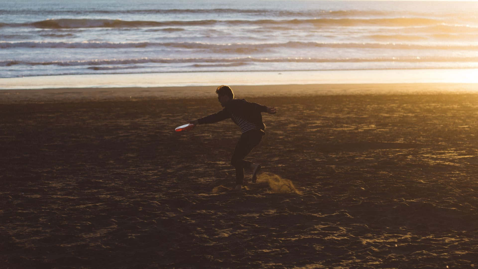 HD Frisbee 桌布 壁紙 — 一個剪影人物在金色日落時分的沙灘上擲飛盤，海浪翻滾而來。