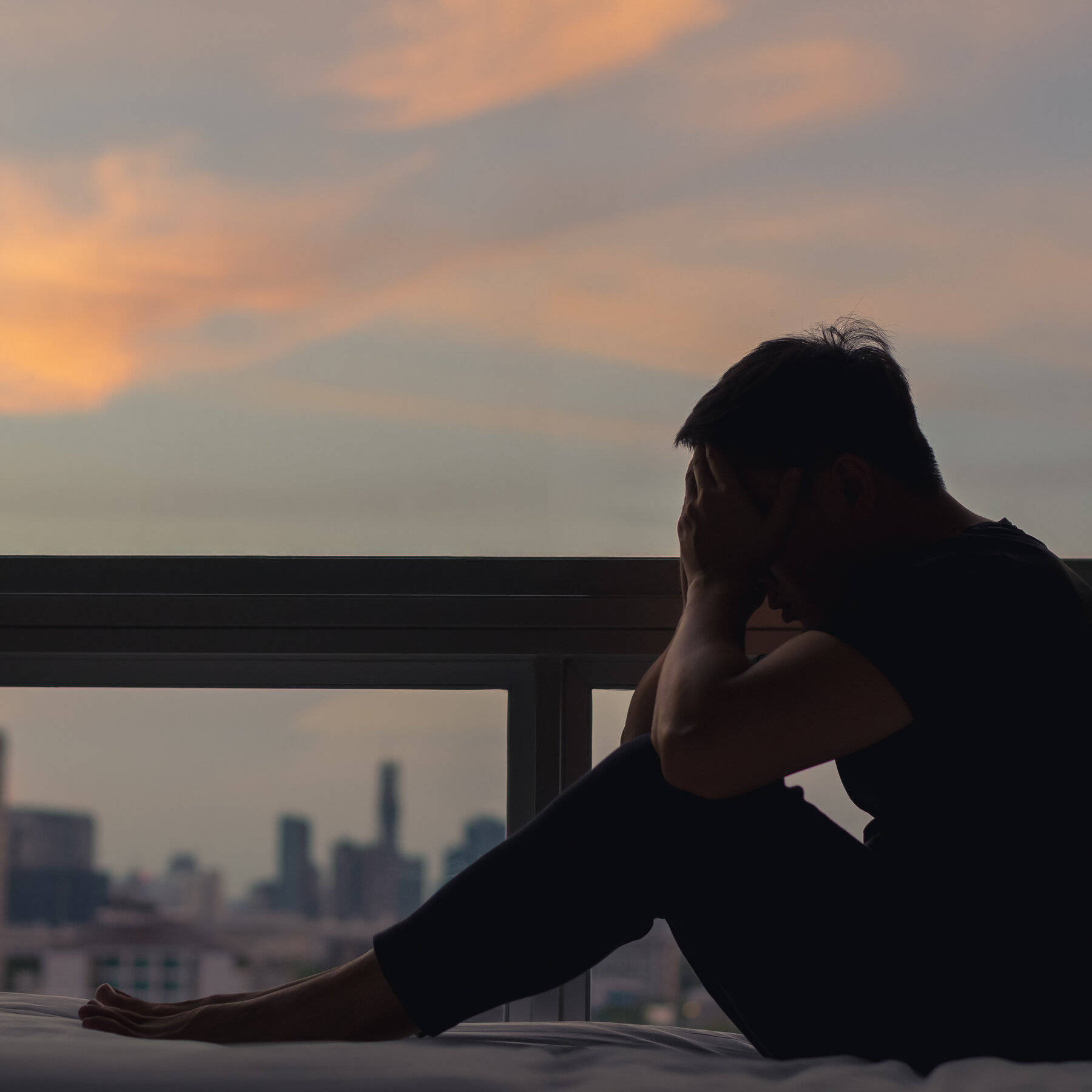 攝影 桌布 壁紙 — 一個剪影人物坐在陽台的床上，夕陽西下，雙手掩面，俯瞰朦朧的城市天際線。