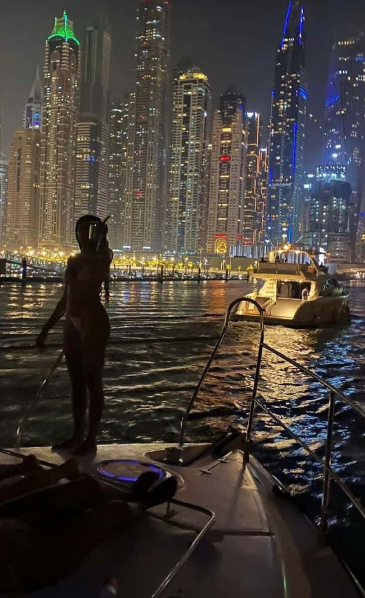 桌布 壁紙 — 一個剪影人物夜晚站在遊艇上，凝視著水中倒映的璀璨杜拜天際線。