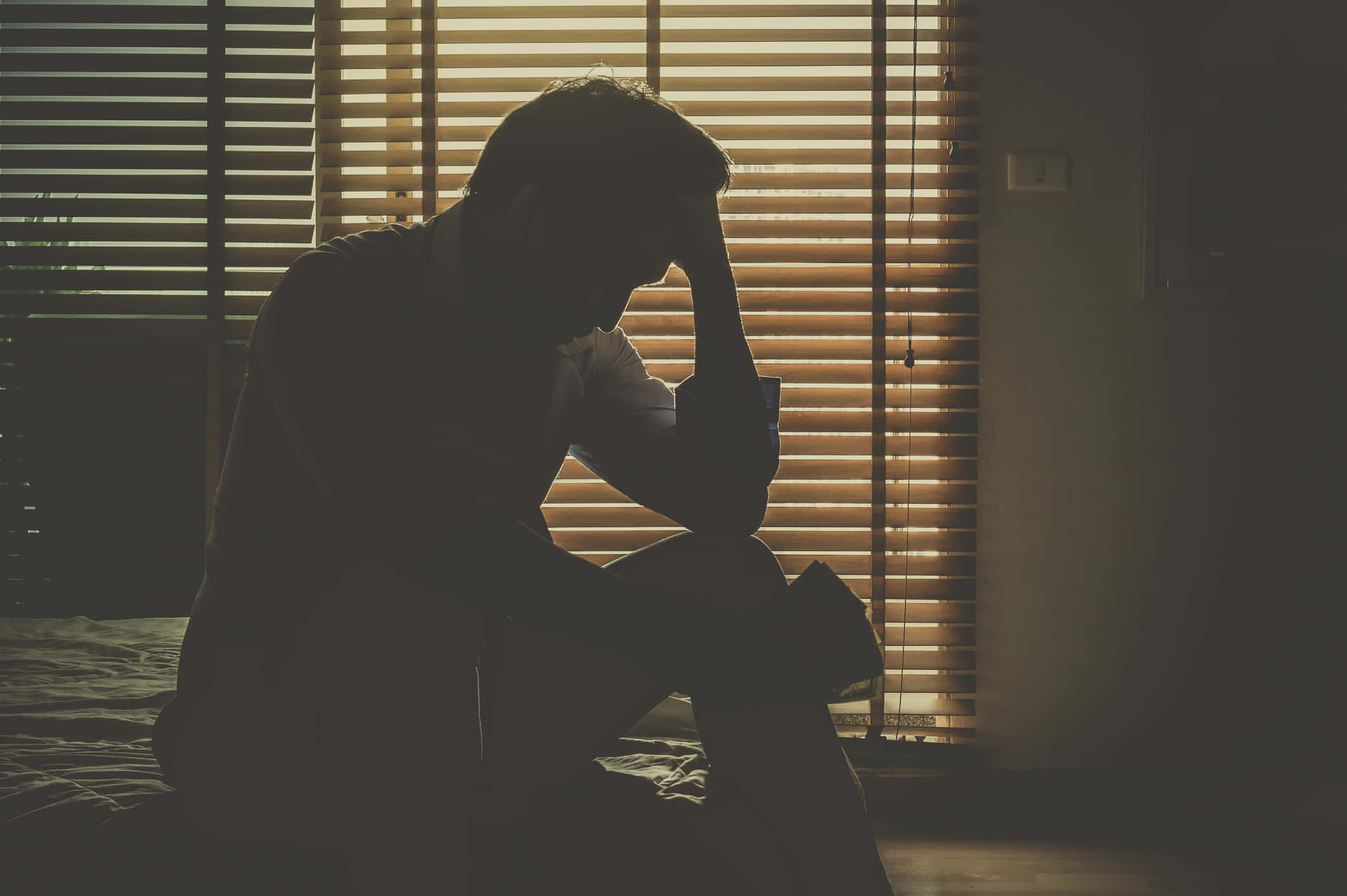 Depressed 桌布 壁紙 — 一個剪影人物癱坐在床上，手托著頭，沐浴在水平百葉窗透進的溫暖光線中。