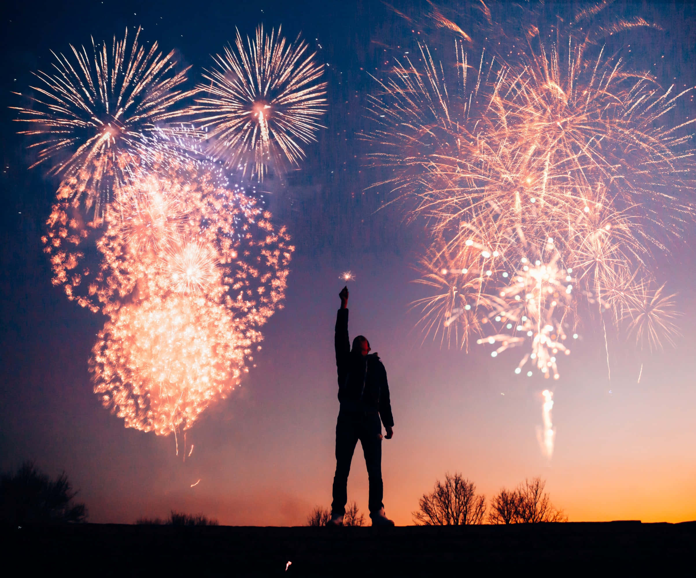 Celebration 桌布 壁紙 — 一個剪影人物站在山丘上高舉仙女棒，而巨大的煙火在暮色天空中綻放。