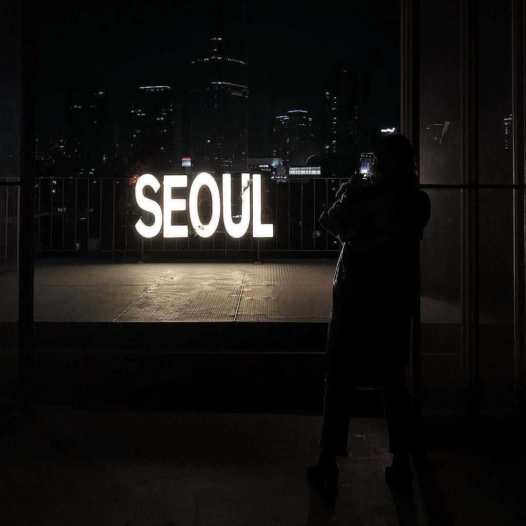 Dark Room 桌布 壁紙 — 一個剪影人物站在發光的「SEOUL」標誌前，夜晚透過窗框可以看到燈火通明的摩天大樓天際線。