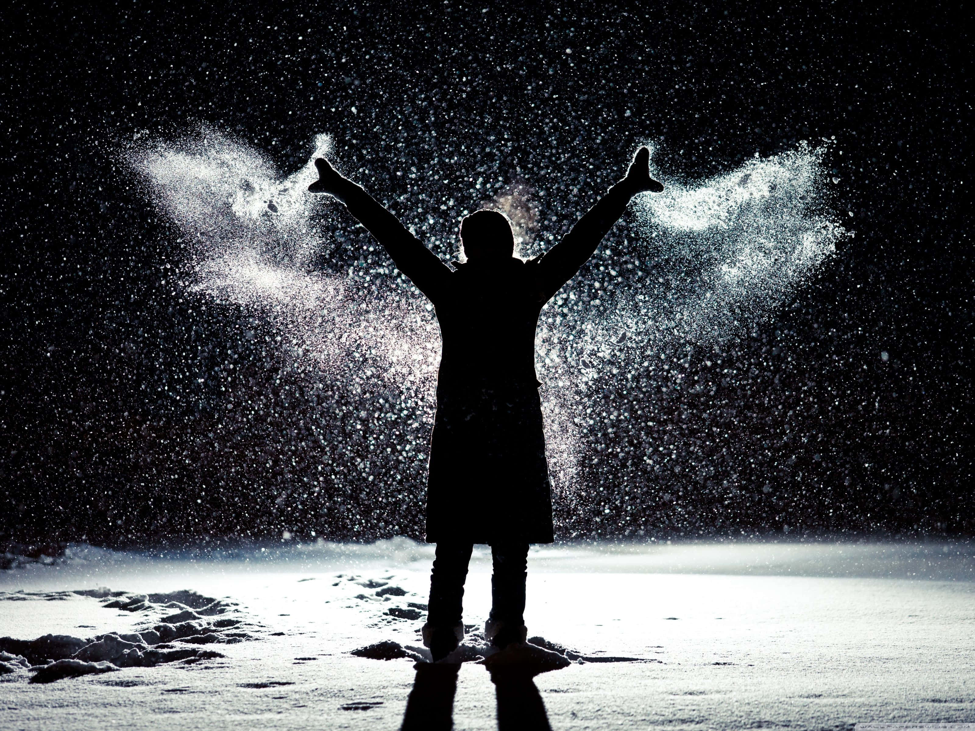 Snow Angel 桌布 壁紙 — 一個剪影人物站在雪灘上，雙臂張開，雪花從他們手中爆炸，映襯著漆黑的夜空。