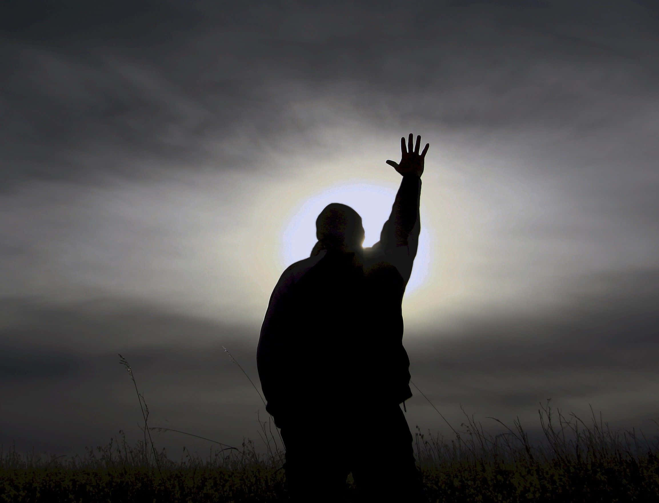 Praying 桌布 壁紙 — 一個剪影人物舉起一隻手，朝向一片草地上方雲層中閃耀的明亮太陽。